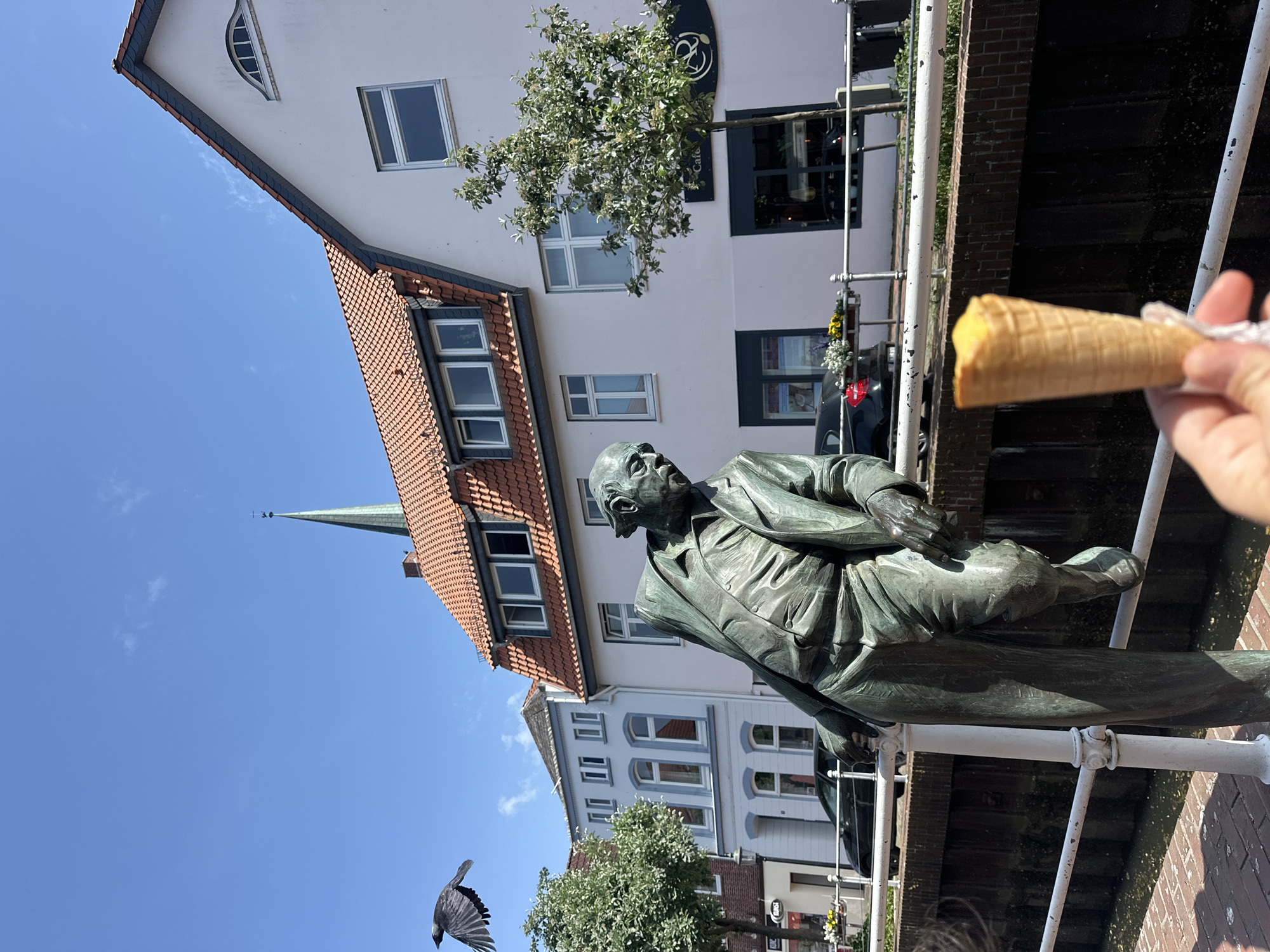 The image shows a bronze statue of a man wearing a suit, leaning against a white railing next to a canal in Buxtehude, Germany. A partly eaten ice cream cone is held in the upper right corner of the frame. The statue is set against a backdrop of buildings with red-tiled roofs and white facades, and a church steeple is visible in the distance. A black bird is flying in the lower left corner of the frame.