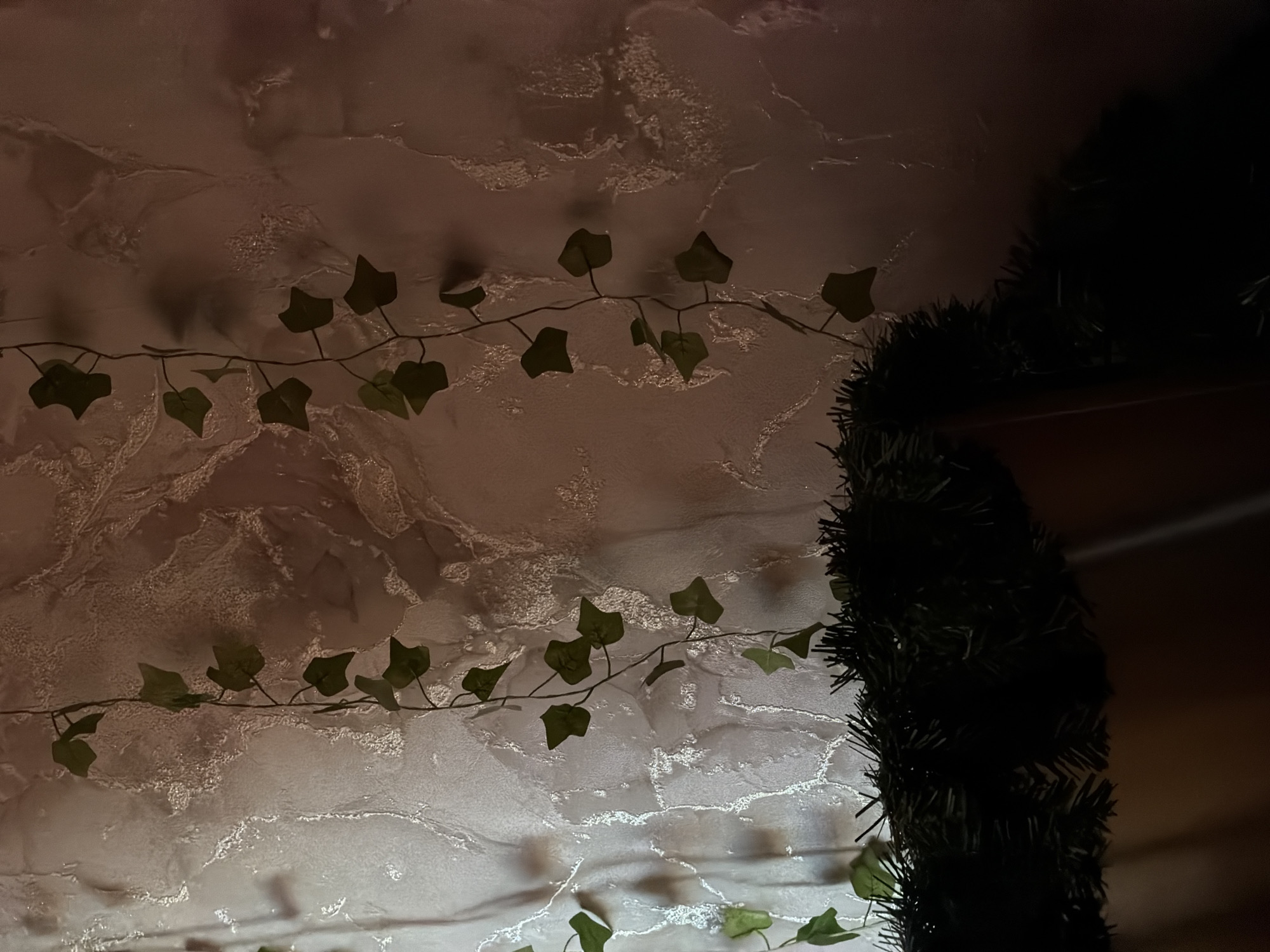 The scene depicts an indoor wall adorned with artificial foliage, located in Perm, Russia. The wall itself features a light-colored, textured surface with an irregular, shimmering pattern, suggesting either decorative wallpaper or plasterwork. Multiple strands of artificial ivy, characterized by dark green, heart-shaped leaves, are suspended vertically against this backdrop. In the lower portion of the frame, a dense segment of dark green artificial evergreen garland or similar decorative foliage is horizontally positioned, largely appearing as a silhouette due to the dim, ambient lighting. No individuals, actions, or specific interactions are discernible within the static scene.