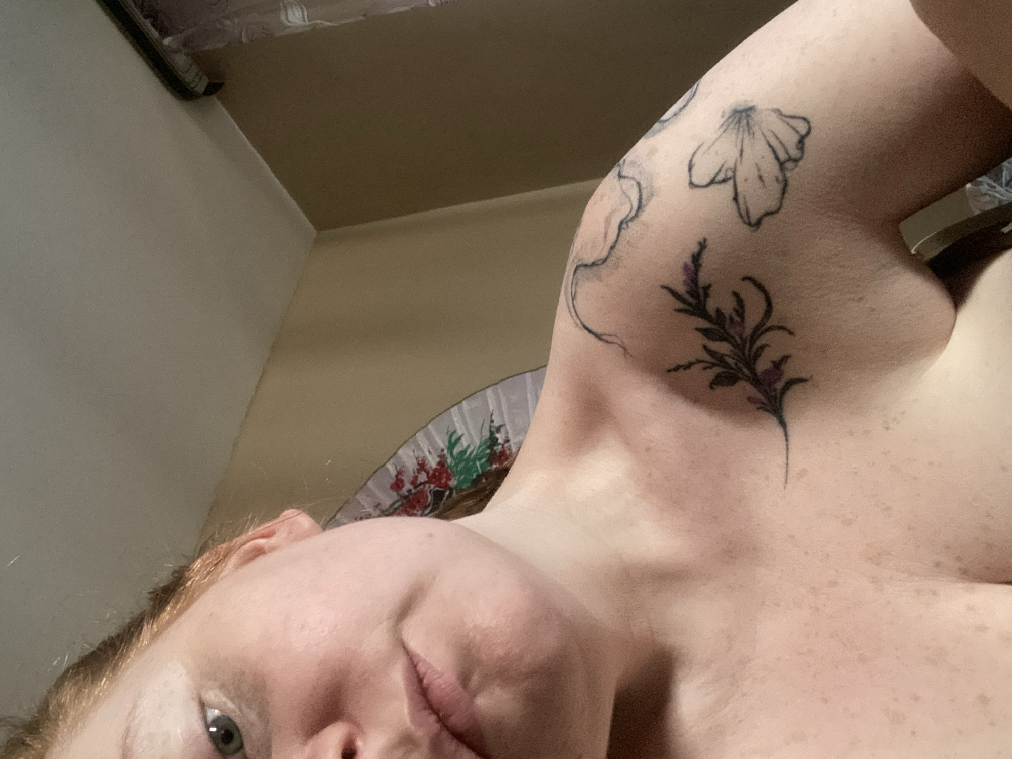 A close-up shot from a low angle captures a portion of a person's upper body. The person has fair skin, with numerous freckles visible on the chest and collarbone. A small part of the person’s face is visible, including the left eye and part of the nose and mouth. The person has reddish-blonde hair. Tattoos are visible on the upper left arm, including flower and plant designs in black ink, with some purple ink highlighting one of the plant designs. A decorative fan is partially visible in the background. The background includes walls and a ceiling, all with a light-colored finish. There is no information suggesting that the photograph was taken in Yekaterinburg, Russia.
