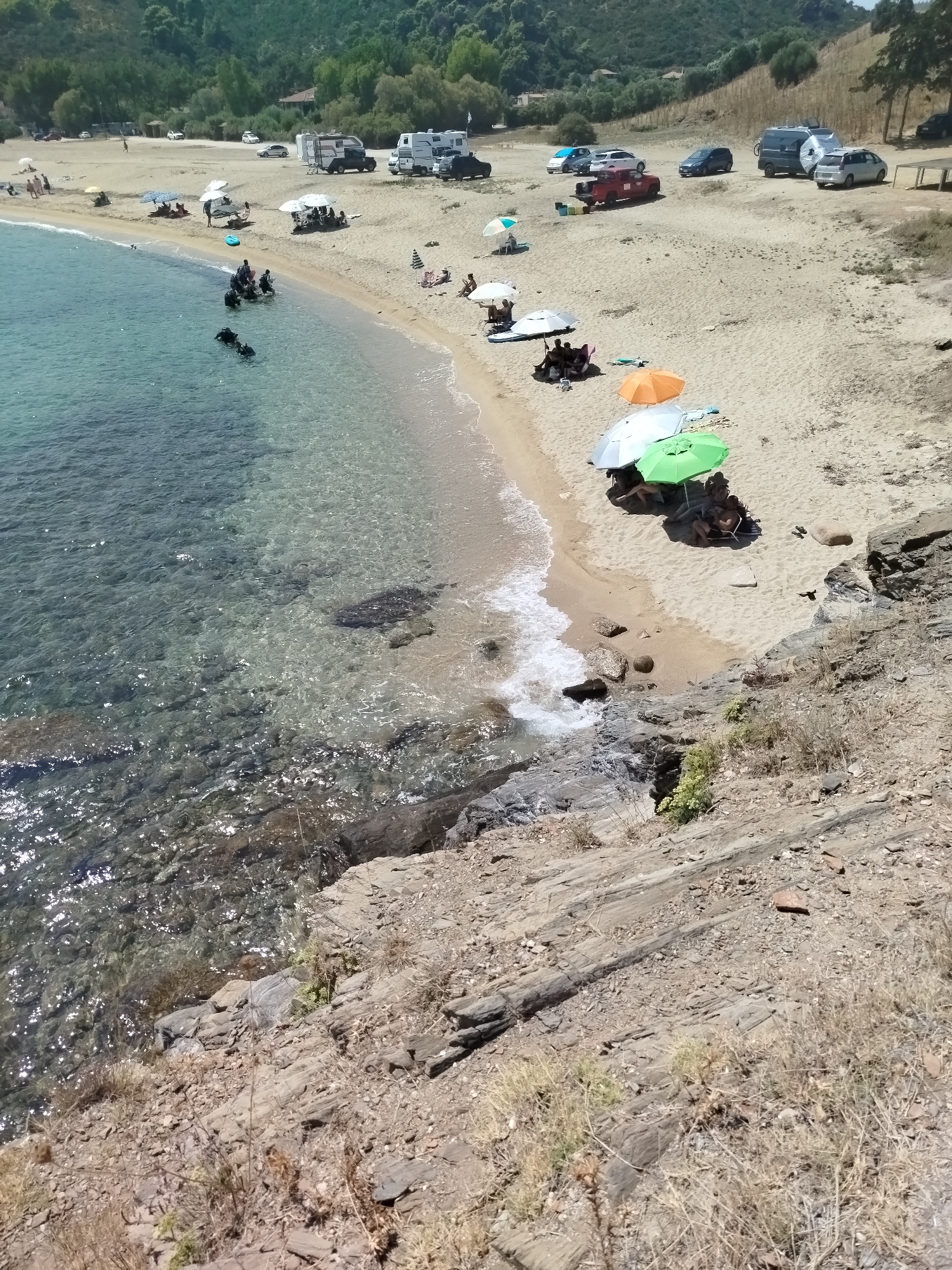 The image depicts a crowded beach scene in Sithonia Municipal Unit, Greece. The sandy beach is bordered by clear, shallow turquoise water on the left and a wooded hillside on the right. Several parked vehicles, including cars, a camper van, and a truck, are visible on the upper part of the beach. Beach umbrellas, chairs, and towels are scattered across the sand, indicating recreational activity. A group of people, some wearing diving gear, are in the shallow water near the shore. Others are seated under umbrellas, relaxing on the sand. The foreground features rocky terrain with dry vegetation, suggesting a natural, undeveloped coastline. The overall atmosphere is one of a busy, sunny day at the beach.