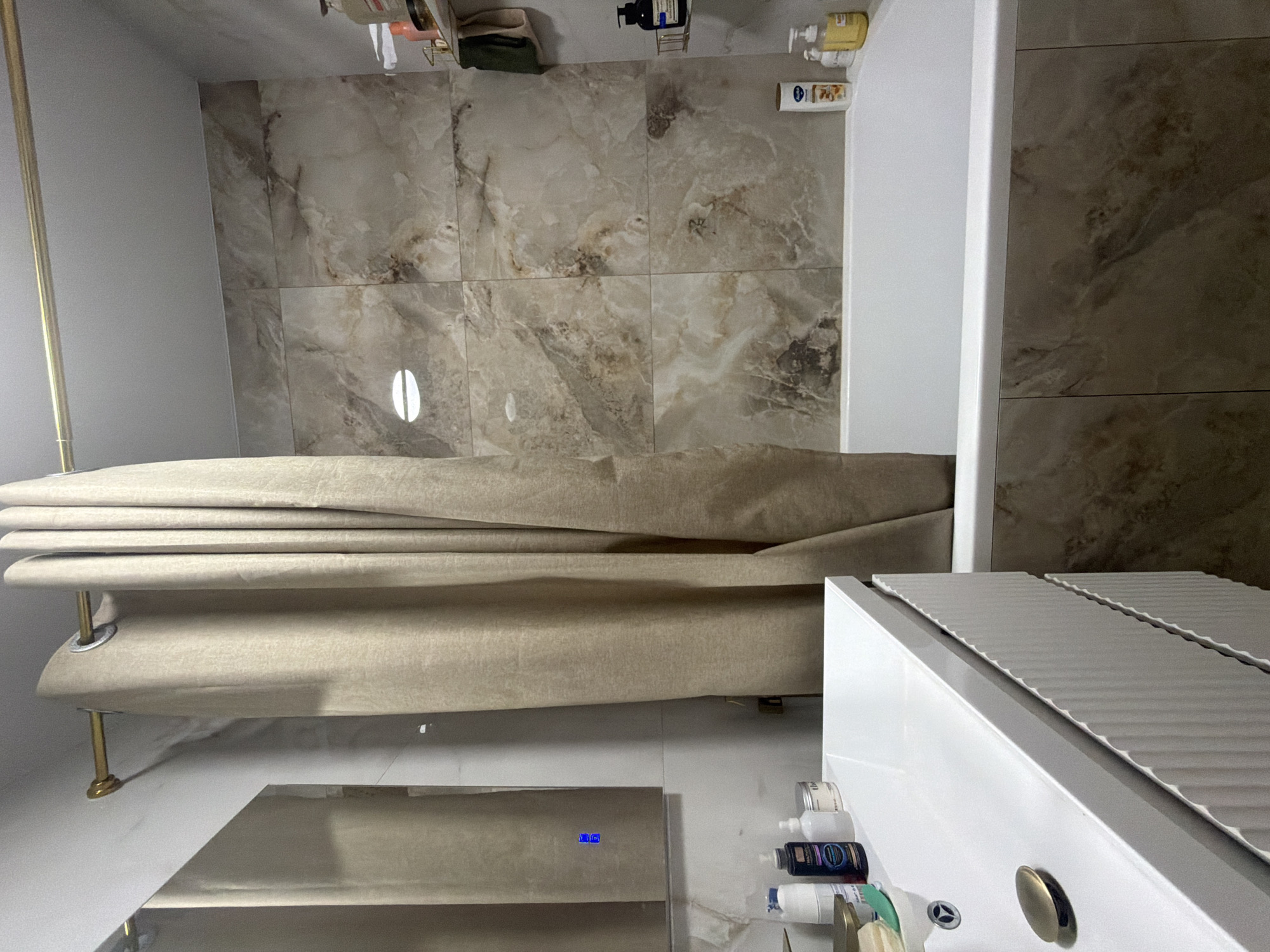 The image shows a modern bathroom with a bathtub, sink, and a shower curtain. The walls are tiled with a marble-like pattern. There are several bottles of toiletries on a shelf above the bathtub. The sink is white and has a gold faucet. The overall impression is one of cleanliness and luxury. The scene is a bathroom in a residential apartment. The bathroom is clean and well-lit. The bathtub is made of white acrylic. The sink is made of white ceramic. The walls are tiled with a beige marble pattern. The shower curtain is beige. The bathroom contains a number of toiletries, including shampoo, conditioner, and body wash. The scene is a bathroom. The image does not depict any people or actions. There are no discernible elements that would indicate this specific location is in Kazan, Russia.