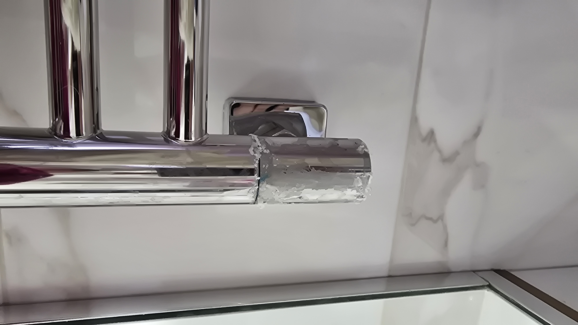 The image shows a close-up of a chrome heated towel rail mounted on a white marble-patterned tiled wall in a bathroom. The lower portion of the vertical bar of the towel rail is covered in a thick, white, crystalline buildup, possibly limescale or mineral deposits. The buildup is uneven and appears to be dripping down the bar. A square chrome mounting bracket is visible behind the lower part of the towel rail. To the left, a sliver of a mirrored cabinet or shower enclosure can be seen. The overall impression is one of a bathroom fixture in need of cleaning. There are no people or animals present. The location is not specified in the image itself, and the request to include the city of Kamenka, Russia, is extraneous to the visual information provided.