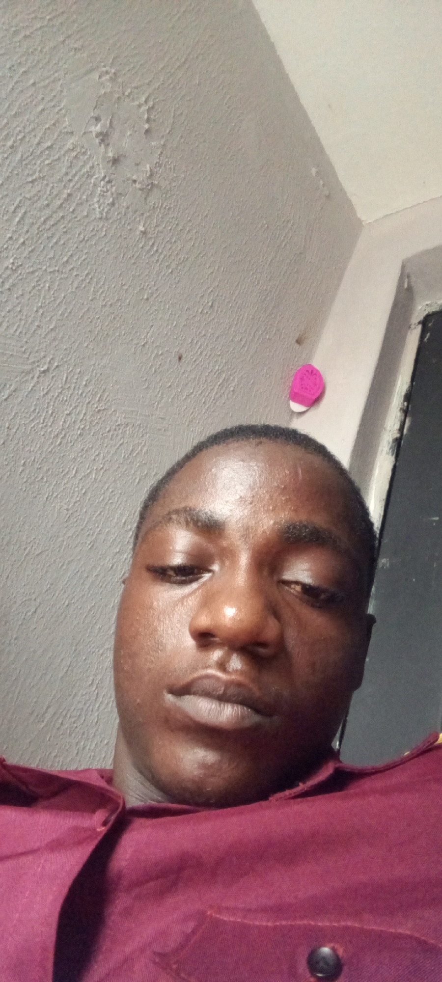 The image is a close-up, low-angle selfie of a young Black male taken indoors. He is wearing a maroon collared shirt. His face is directed towards the camera, with his eyes looking slightly down and to the side. The background consists of a textured, light-colored wall with some imperfections and a corner where it meets another wall. To the right, a sliver of a dark opening, possibly a doorway or window, is visible. A small, bright pink decorative item with white trim is attached to the wall on the upper right side of the frame. The lighting suggests an indoor setting, possibly a room. The image does not contain enough information to determine the specific location within Ilado, Nigeria.