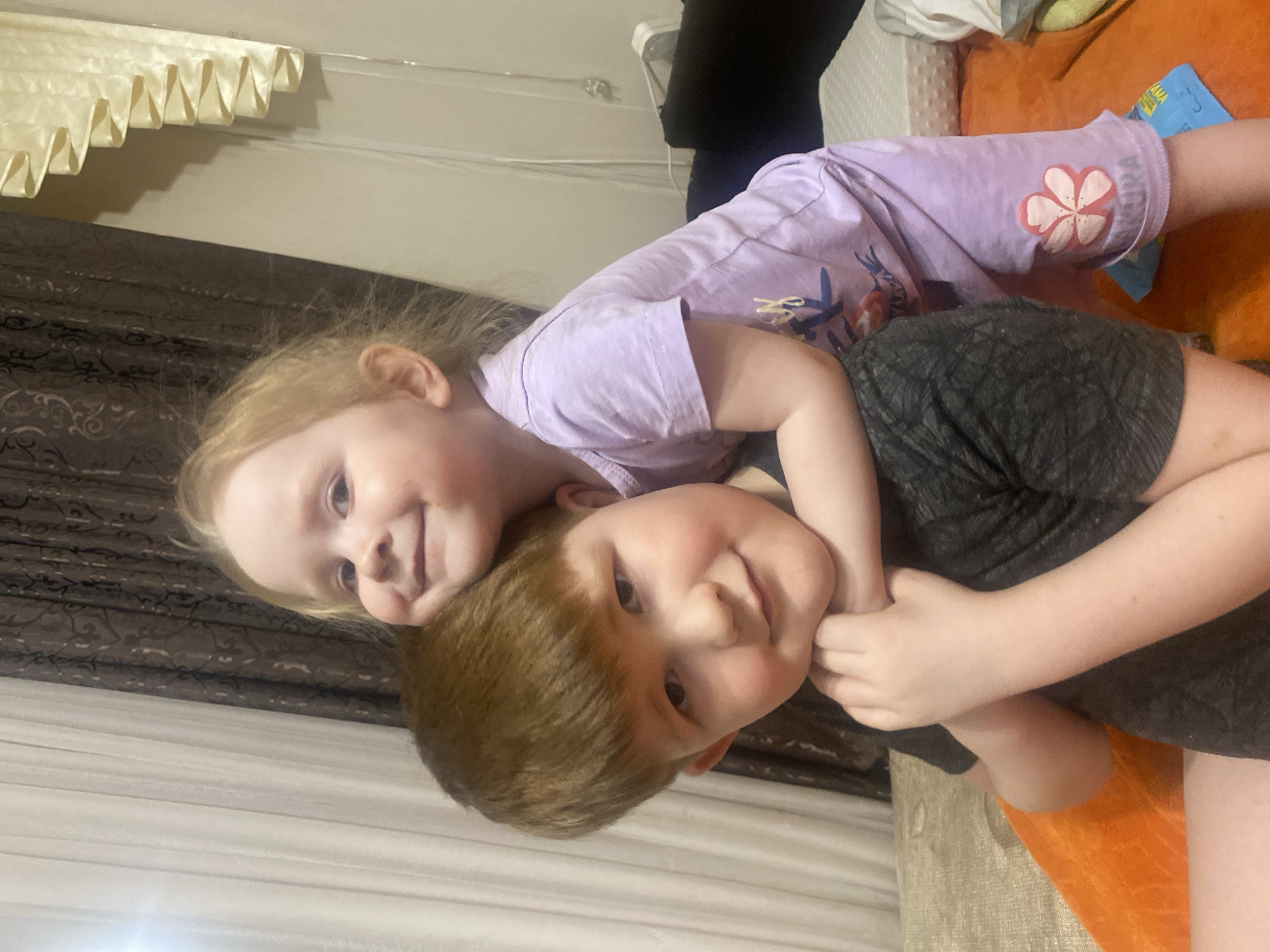 Two young children are interacting. One child, with blonde hair, is lying atop the other child, who has brown hair. The blonde child is wearing a lavender t-shirt with a floral design. The brown-haired child is wearing a dark gray shirt. They are lying on an orange surface, likely a bed. The background includes curtains and a patterned wall covering. The location is Chaikovsky, Russia.