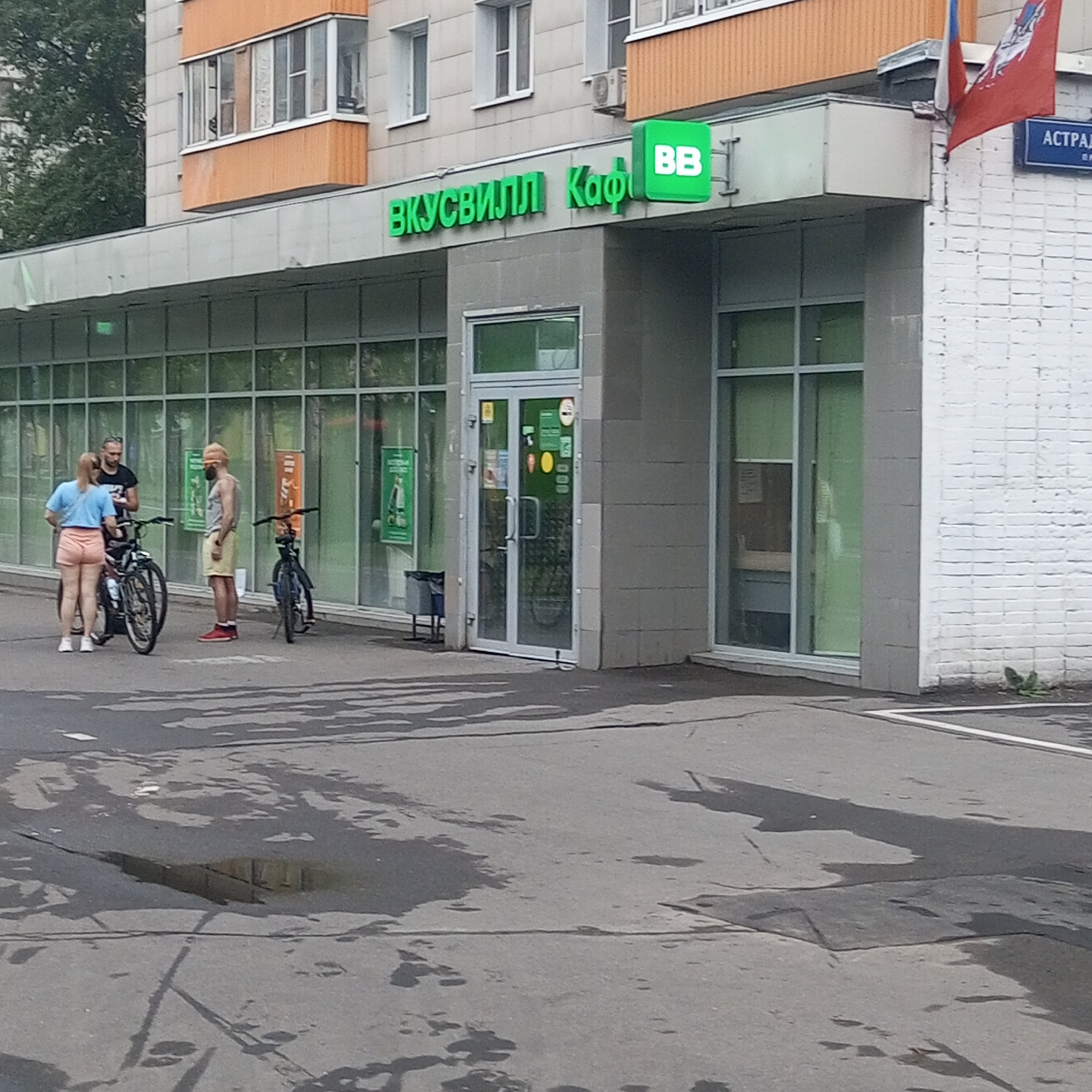 The image shows the facade of a "VkusVill" grocery store and cafe in Moscow, Russia. A green sign with white lettering reads "ВКУСВИЛЛ Кафе" and a separate green square sign has "BB" in white letters. Three people are standing outside the store near two bicycles. One person, a woman wearing a blue t-shirt and peach shorts, is facing the camera and standing next to a black bicycle. Another person, a man wearing a black t-shirt, is also facing the camera, standing behind the woman and her bicycle. A third person, a man wearing a light-colored tank top and yellow shorts, is standing to the right of the other two, facing away from the camera, next to a blue bicycle. The store has large glass windows and a double glass door entrance. A street sign with blue lettering reading "АСТРАХАНСКАЯ" is visible on the right side of the building, attached to a white brick wall. The ground in front of the store is wet and reflects the surrounding environment.