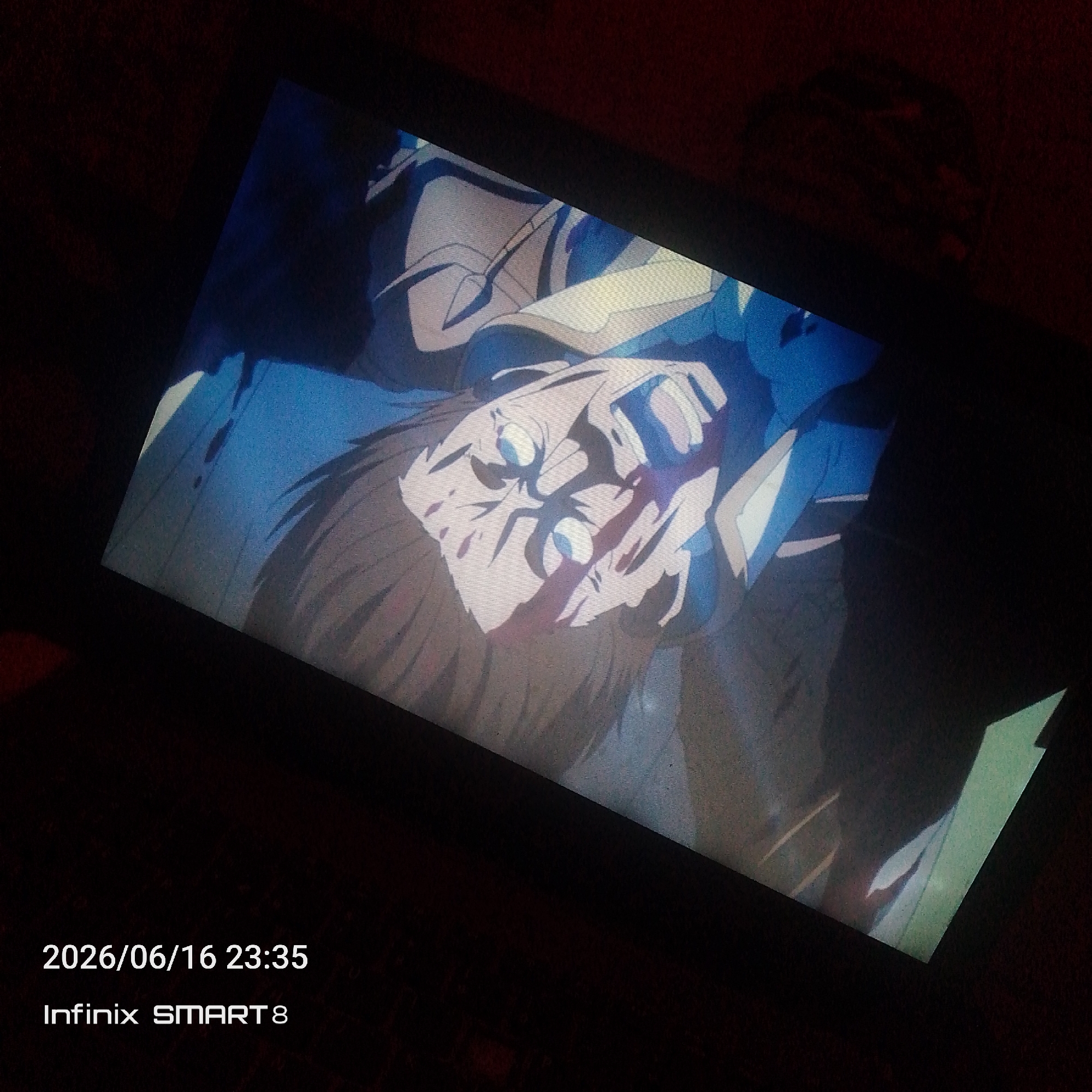A close-up, angled photograph taken in low light conditions shows a portion of a digital screen displaying an animated scene. The animation depicts a close-up of a character's face, which appears to be a man with fair skin, prominent cheekbones, and light blue eyes. He has dark hair and is wearing what looks like armored clothing in shades of blue and white. The overall color palette of the animation is dominated by cool tones of blue, white, and gray, with some darker shadows. The screen is tilted, and the edges of the device are visible, along with a dark, textured background that appears to be fabric, possibly a sofa or chair. In the lower left corner of the screen, a timestamp "2026/06/16 23:35" and device information "Infinix SMART 8" are overlaid. There is no discernible location within the city of Jos, Nigeria.