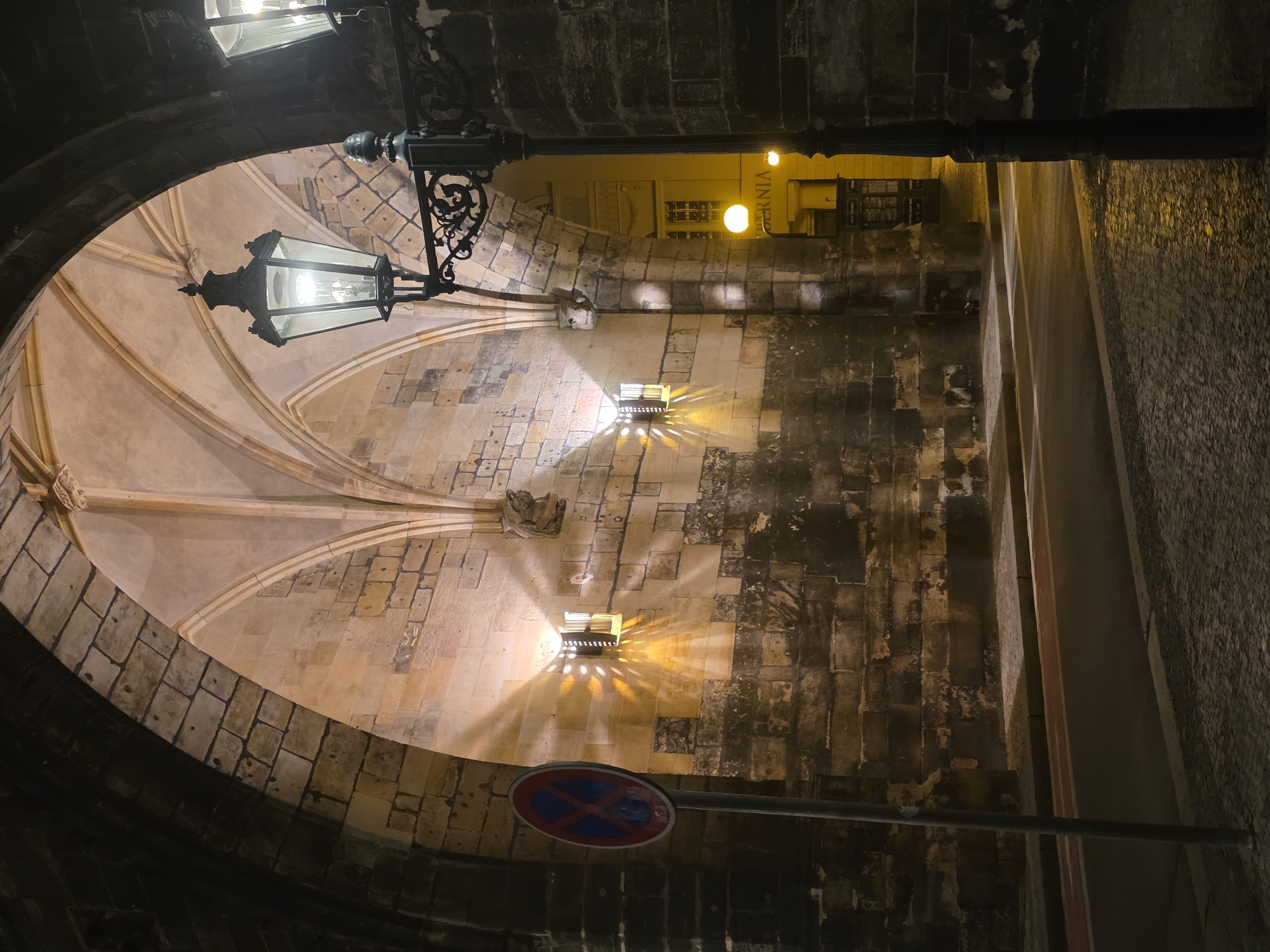 The image captures a nighttime scene looking up into an ornate, arched stone passage or underpass. A large, dark metal lantern hangs prominently from the archway in the foreground, its bright light illuminating the ribbed vaulting of the ceiling, which displays a warm, light beige tone. The passage walls are constructed from rough, irregular stone blocks, showing signs of age.
Beyond the main arch, to the right, a street or building facade is visible, bathed in a warm yellow light from several glowing orbs and other fixtures. On the right wall of the passage, a smaller, square-shaped wall-mounted light casts a distinctive starburst pattern of illumination onto the stone surface below it.
In the lower right portion of the image, a circular red and blue "no stopping" road sign is attached to a black pole. The artificial lighting, combined with the stone architecture, creates a historic and atmospheric ambiance. The time of day is clearly night, as indicated by the deep shadows and dependence on the various light sources. Part of a word, "VERNIA" or "MODERNIA" (partially visible and inverted in the original image but clearer in crops), can be faintly discerned on a sign in the brightly lit background area. No people or activities are present. The architectural details, especially the vaulted ceiling, suggest an old European urban setting.