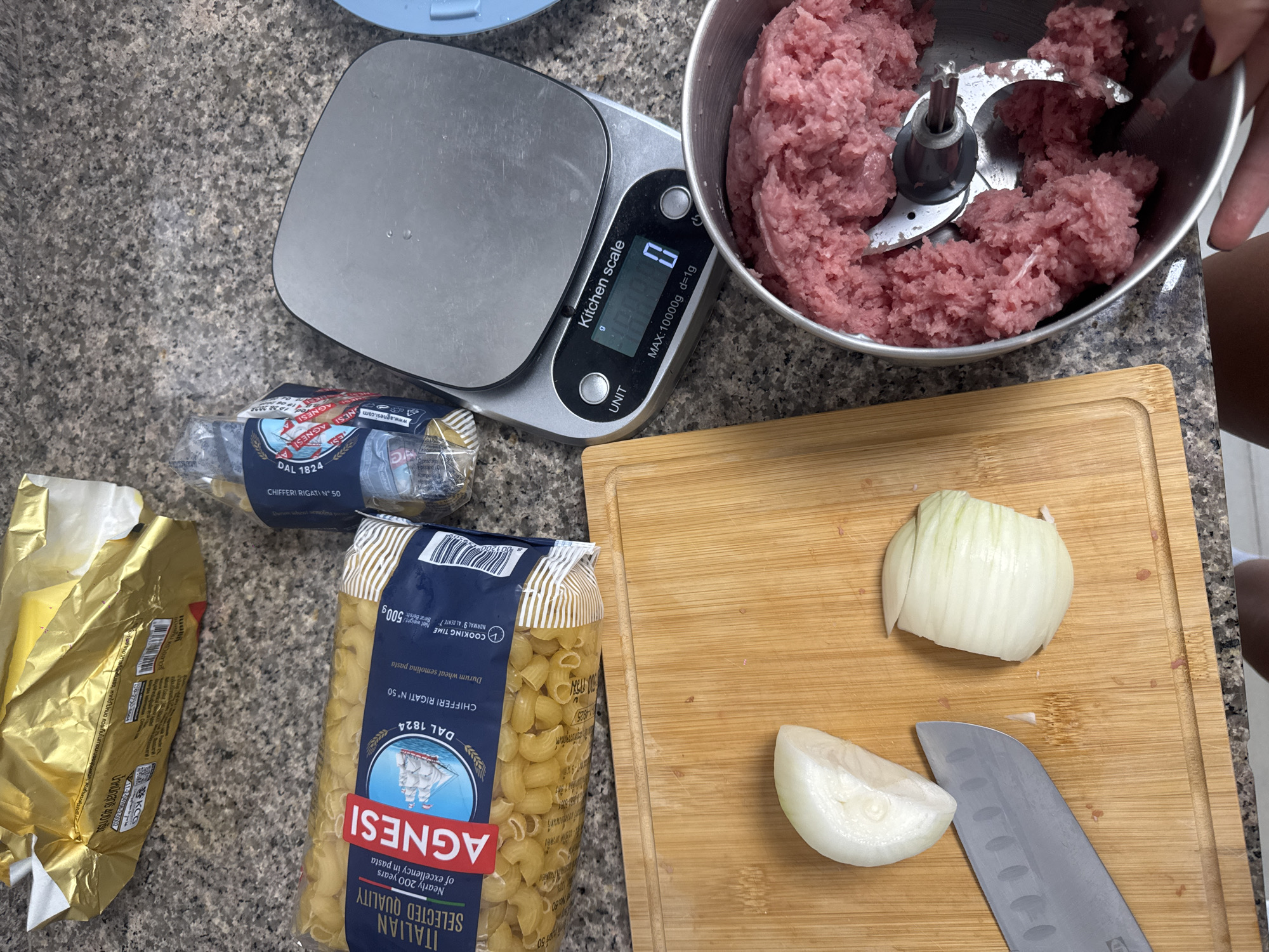 The image depicts a countertop scene during food preparation, but there's no evidence to suggest it's located in Chalong, Thailand.
On the counter, there's a kitchen scale displaying "0 g," a stainless steel bowl filled with ground meat and a food processor blade. Next to the scale, there's a package of Agnesi brand "Chifferi Rigati N° 50" pasta and another package of pasta and butter.
A wooden cutting board with a partially chopped white onion and a kitchen knife are also visible. A hand is seen holding the bowl of ground meat. The counter appears to be a gray stone surface.