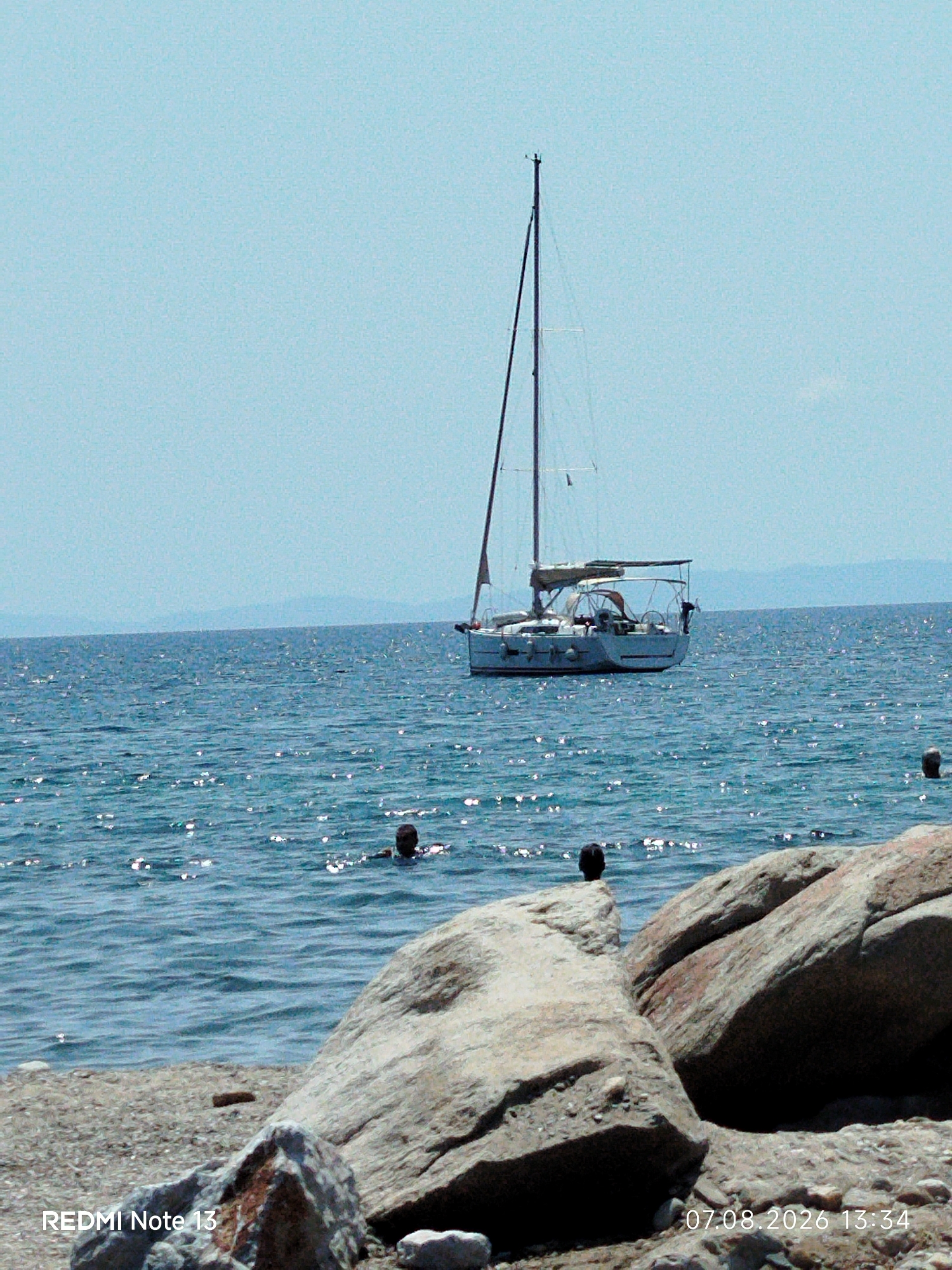 A sailboat is moored in the clear blue waters off the coast of Sithonia, Greece. The boat is white with a tall mast and a dark sail. Several people are visible swimming in the water, their heads bobbing above the surface. In the foreground, large rocks line the shore, with a beach of pebbles stretching beyond. The sky is a pale blue, and the water is calm and reflects the sunlight. The timestamp "07.08.2026 13:34" and the text "REDMI Note 13" are overlaid on the bottom of the image.