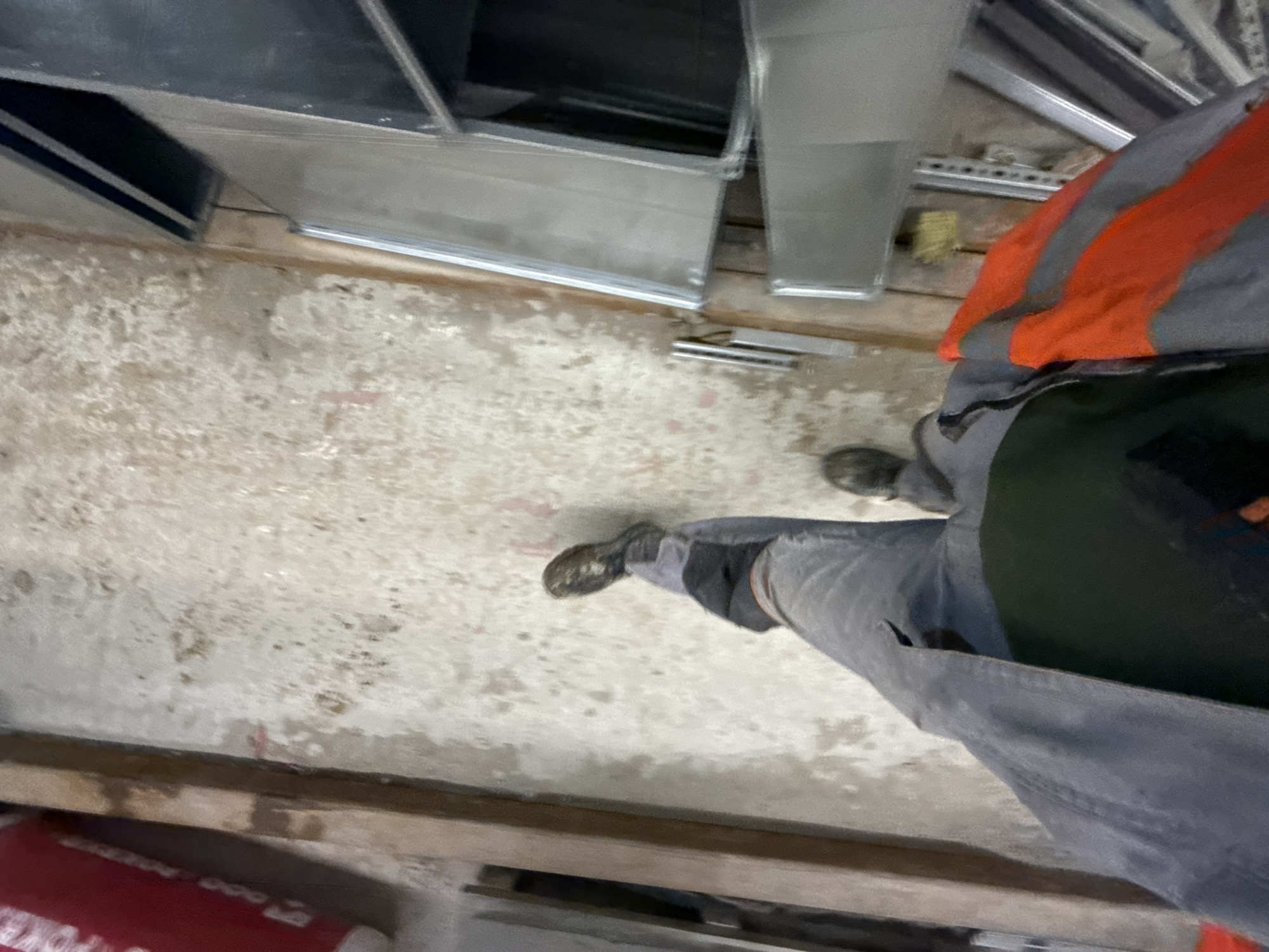 The image shows a first-person perspective of someone walking on a concrete floor. The individual is wearing work boots, gray pants, and a dark jacket over a green shirt. A bright orange safety vest with gray reflective stripes is visible on the right side of the frame. The environment appears to be an indoor construction or industrial site, with some wooden beams and metal framing structures visible. The floor is rough and stained. There is a red cylindrical object with white lettering on the left side of the image. The image is blurred and lacks sufficient detail to determine the specific location within Moscow, Russia, or the exact nature of the work being performed.