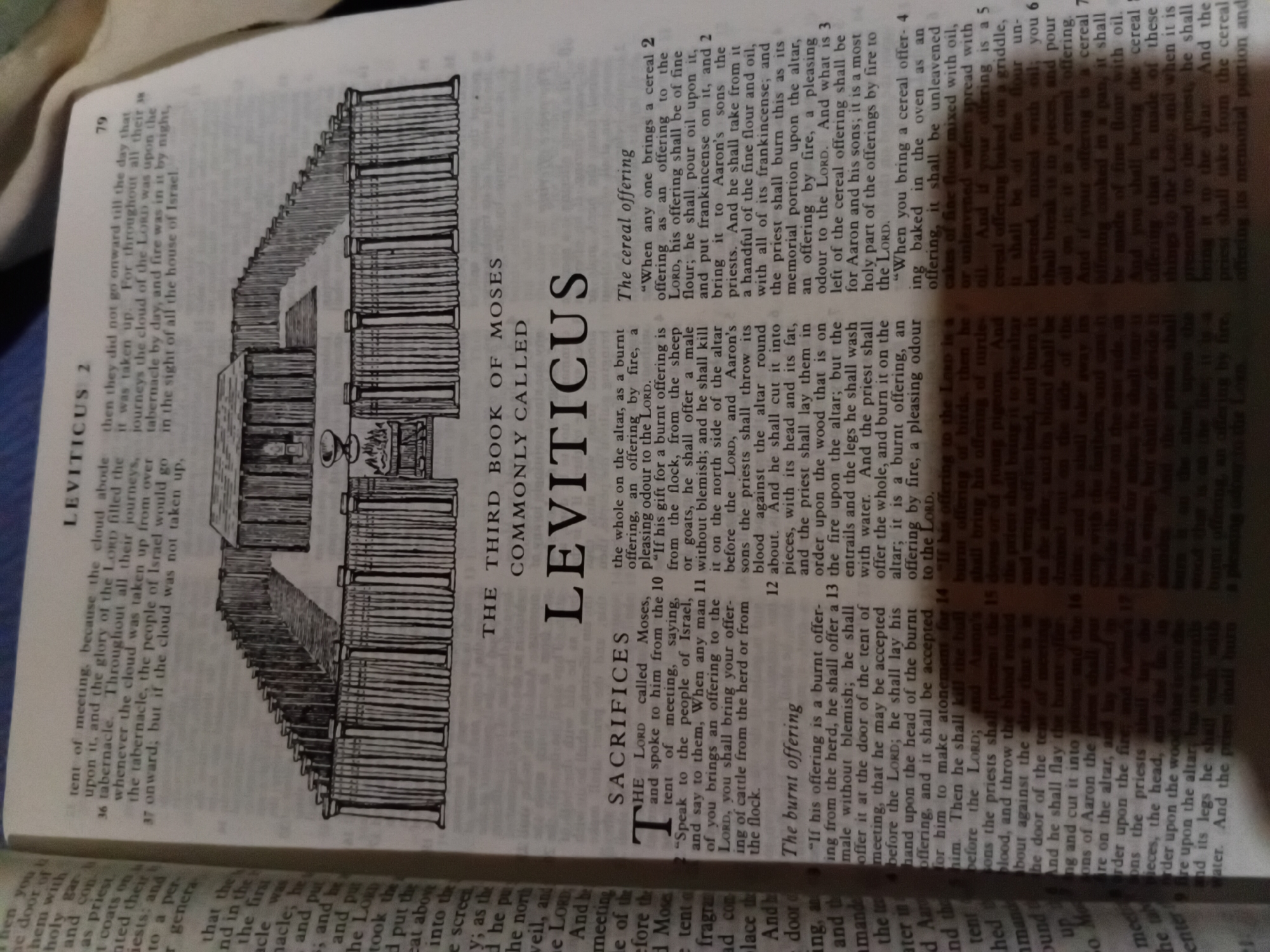 This is a photograph of an open Bible, displaying text from the book of Leviticus, alongside an illustration. The text is in English, with the chapter title "Leviticus 2" visible at the top of the page, along with the page number "79". Below the chapter title, a subheading reads "THE THIRD BOOK OF MOSES COMMONLY CALLED LEVITICUS".
The page is divided into two columns of text, with a large black and white illustration positioned between the upper sections of the columns. The illustration depicts a walled enclosure with a central structure, resembling ancient religious architecture.
The text on the left side of the page begins with the subheading "SACRIFICES" and details various offerings, including "The burnt offering". The text on the right side of the page includes subheadings for "The cereal offering".
The photograph is taken from a slightly elevated angle, with the book lying flat. The lighting appears to be natural, with some shadows cast on the pages. The background is blurred, suggesting the book is the primary subject of the photograph. There is no indication in the image or the provided text of the city of Nkpor, Nigeria, or any forensic details pertaining to a crime scene.