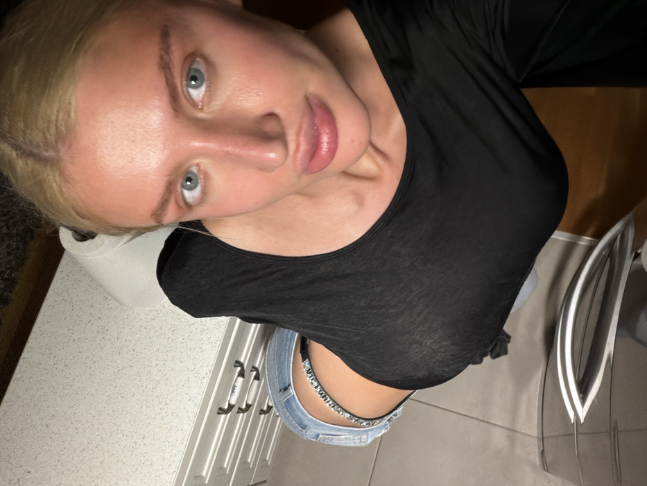 A high-angle selfie captures a fair-skinned individual with blonde hair pulled back, light blue eyes, and a visible reddish complexion/tan on the face. The individual wears a form-fitting, long-sleeved black top and light blue denim jeans. A thong-style undergarment with a visible sparkling or rhinestone-embellished waistband is exposed above the jeans. The scene is an indoor domestic setting. In the background, a white, speckled countertop is visible, upon which a roll of white paper towels rests. Below the countertop, light-colored cabinets with dark horizontal bar pull handles are partially visible. In the lower foreground, a dark, reflective surface with a chrome-like handle is present. The floor appears to be a light-toned, hard surface. The precise location within the city of Budva, Montenegro, cannot be determined from the visual information provided in the image.