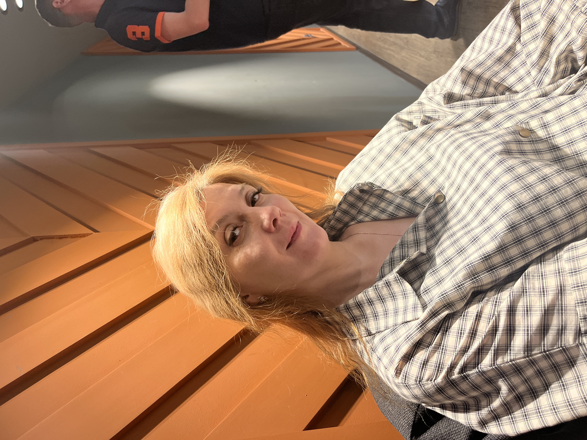 A woman with blonde hair is lying on an orange, wooden structure. She is wearing a plaid shirt. In the background, a person is wearing a black shirt with orange accents. The scene appears to be indoors. Izhevsk, Russia is not identifiable in this image.