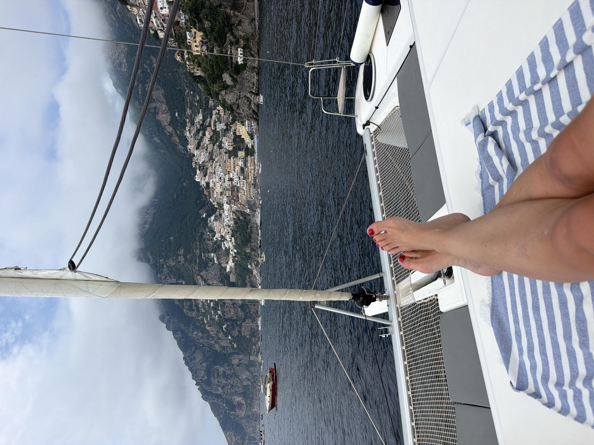 The image depicts a view from a boat, possibly a sailboat or catamaran, towards the town of Positano on the Amalfi Coast in Italy. Positano's multi-colored buildings are visible, clustered along the steep hillside that plunges into the sea. The surrounding landscape features rocky cliffs and green vegetation, partially obscured by low-hanging clouds or mist. Part of the boat is visible, including the netting on the deck and rigging. The legs and feet of a person are visible on the deck, painted with red nail polish and a striped towel is present. In the water, a small boat can be seen anchored or moving.