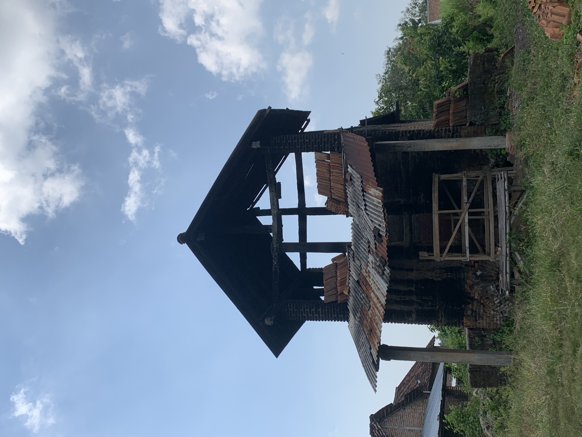 The image depicts an abandoned brick kiln structure in Gulun, Indonesia. The kiln, constructed primarily of dark brick, is in a state of disrepair. Its roof is partially covered by corrugated metal sheeting, showing signs of rust. Several stacks of terracotta tiles are visible, some piled on the structure and others on the ground. The surrounding environment is overgrown with tall grass and green foliage, with a few other buildings in the background that also appear to be made of brick. The sky is clear blue with scattered white clouds. There are no people or animals visible in the image.