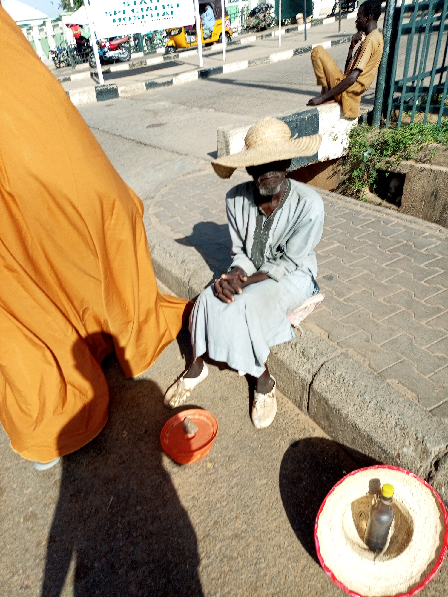 The image shows a street scene in Arawa, Nigeria, during the day. Two men are visible. One man, wearing a large straw hat and a light gray robe, sits on a raised curb next to a paved walkway. His hands are clasped in his lap, and he appears to be resting. In front of him, on the ground, are an orange pot and a straw hat with a red trim, which contains a dark bottle. A large, bright orange cloth is draped to the left of the frame, partially obscuring the man in the gray robe.
Further back and to the right, a second man sits on the curb, facing away from the camera. He is wearing a light brown outfit and has his legs crossed. He appears to be looking to the right. Behind him, a dark green metal fence runs along the edge of the sidewalk. In the background, there are vehicles, including a yellow tricycle and motorcycles, and a sign that partially reads "SPECIALIST HOSPITAL." The sunlight suggests it is a bright, possibly warm day, indicated by the strong shadows cast. The overall impression is of a busy roadside environment.