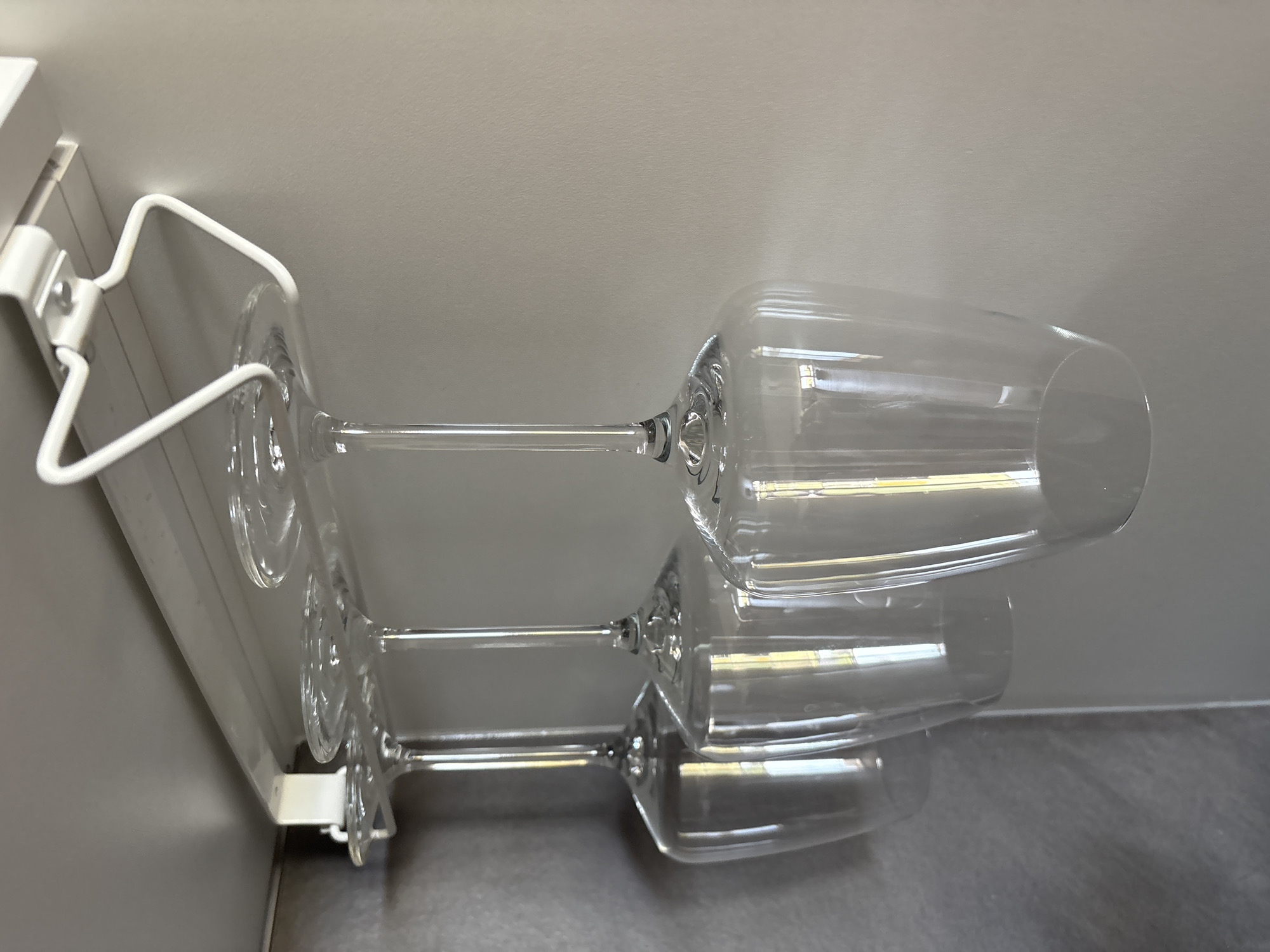 Three clear wine glasses hang upside down from a white rack mounted under a grey shelf. The glasses are positioned in a line, with their stems facing the viewer and their bowls extending downwards. The background consists of a plain, light grey wall. The scene is illuminated by overhead lighting, creating reflections on the glass surfaces. The image appears to be taken indoors, possibly in a kitchen or pantry. The location in Bönnigheim, Germany, is not discernible from the visual information provided in the image.
