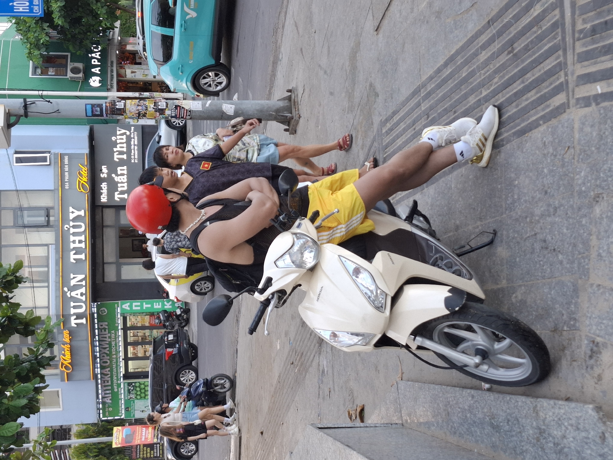 The image shows a street scene in Nha Trang, Vietnam, with several people and a parked scooter. In the foreground, a person wearing yellow shorts and white sneakers is sitting on a scooter, leaning back. Another person, wearing a dark shirt with a red star emblem, is also on the scooter, holding the handlebars. A red helmet rests on the scooter. Behind them, two other people are standing, one wearing a patterned shirt and blue shorts, and the other in a white shirt.
The background features several buildings with signage in Vietnamese and Russian. One prominent sign reads "Khách Sạn TUẤN THỦY" (Tuấn Thủy Hotel) with an address and phone number. Another sign, "АПТЕКА ОРХИДЕЯ" (Orchid Pharmacy) is also visible. A teal-colored car is parked further down the street.
The scene appears to be during the daytime, with ample natural light. The overall atmosphere suggests a casual street environment in an urban setting, possibly a commercial area given the hotel and pharmacy signs. The people seem relaxed, with no indication of urgency or distress. The scooter and the vehicles suggest a mode of transportation common in the area. The presence of diverse signage might indicate a tourist or commercial district.