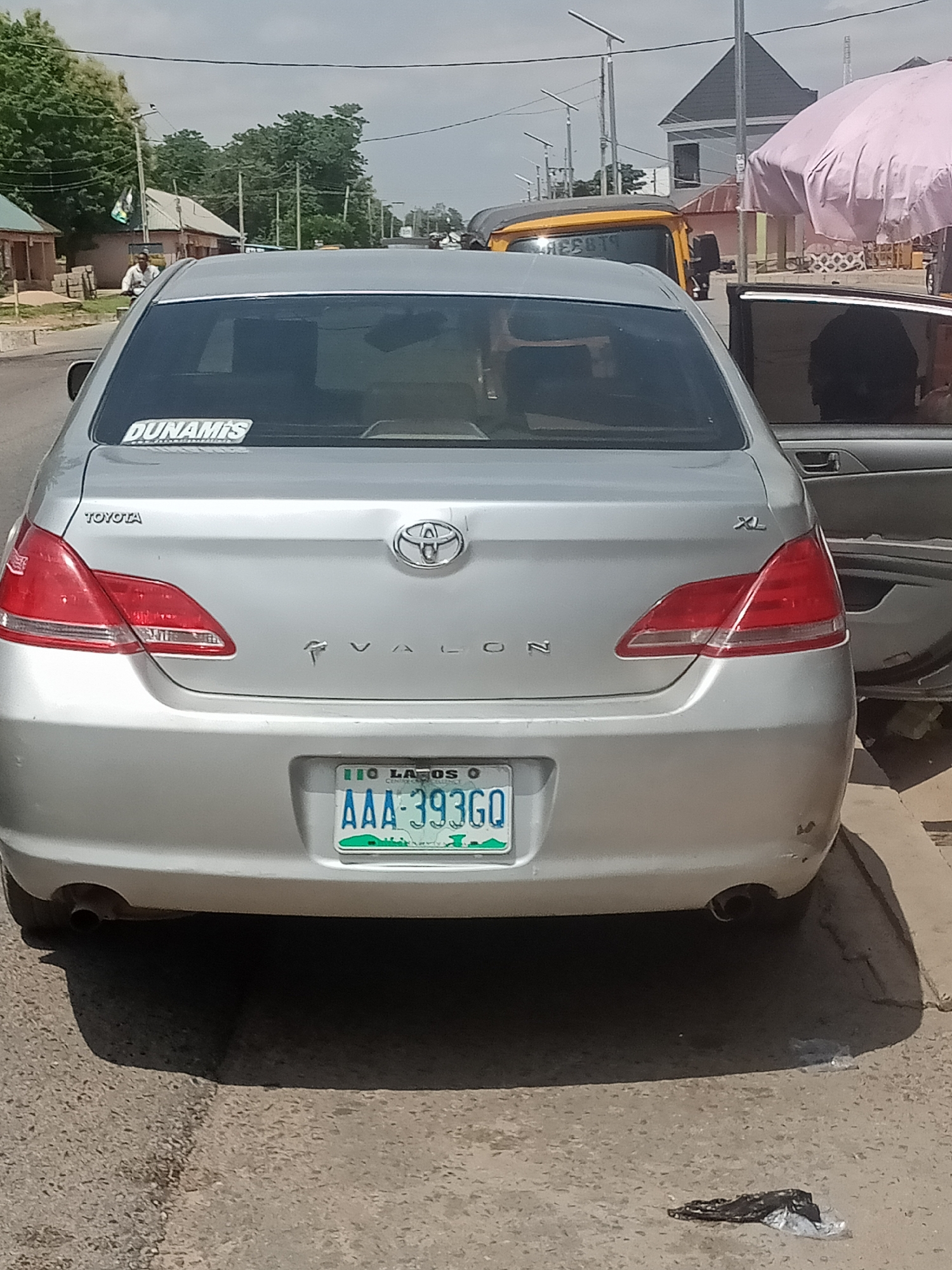 The image shows the rear view of a silver Toyota Avalon sedan parked on a paved road. A "DUNAMIS" sticker is visible on the rear window. The license plate reads "AAA-393GO" and is green, with "LAGOS" printed at the top. The car has two taillights and two exhaust pipes. The passenger side door is ajar. In the background, there are buildings, trees, and a yellow tricycle with the word "REBBYT" visible on its side. A person is seen riding a motorcycle in the distance. The scene appears to be in Jalingo, Nigeria, as indicated by the license plate. The sky is cloudy.