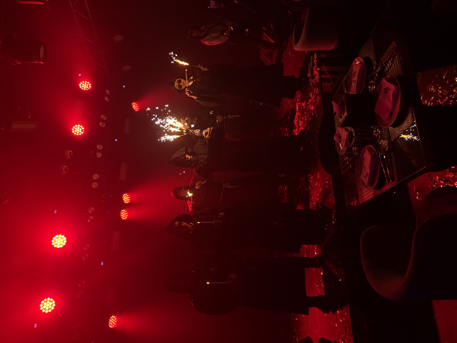A dimly lit room with a red hue showcases a group of individuals dressed in dark robes, possibly hooded, each holding a burning object, possibly candles or sparklers. The lights cast a strong red color across the room, illuminating a few tables set with plates and cutlery. The atmosphere appears theatrical, potentially a performance or celebration.