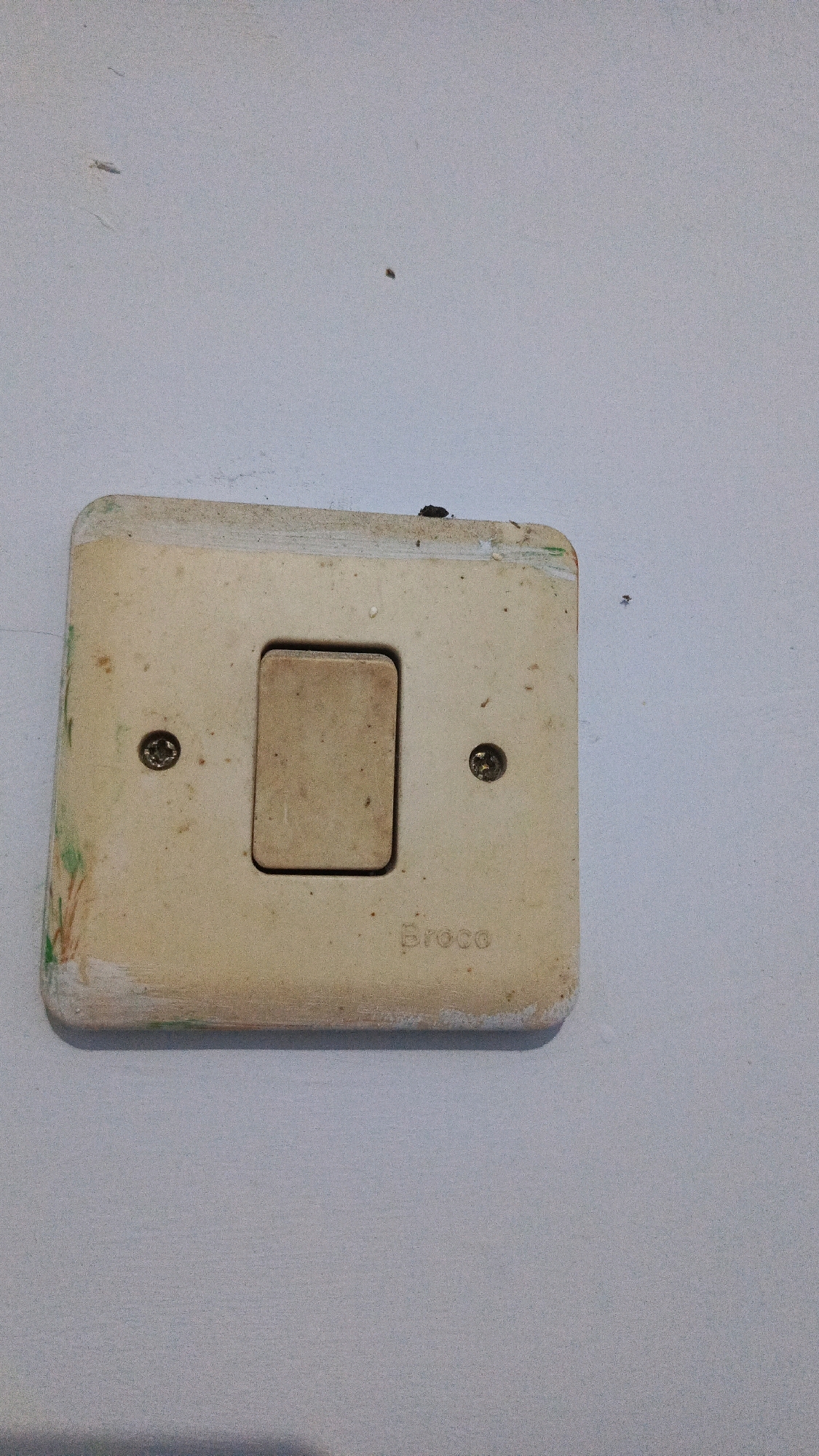 This is a close-up, eye-level, medium shot of a light switch on a white wall. The switch is an older, cream-colored plastic model with a single toggle. It is mounted on a square plate, which has some green and pink marks on its lower left side. Two screws are visible on the plate, one on either side of the toggle. The brand name "Broco" is embossed on the lower right of the plate. The wall has some minor blemishes and a couple of small, dark specks that appear to be insects or debris. The lighting is neutral, with no strong shadows. The image appears to be taken indoors in a residential or commercial setting within the city of Babadan, Indonesia.