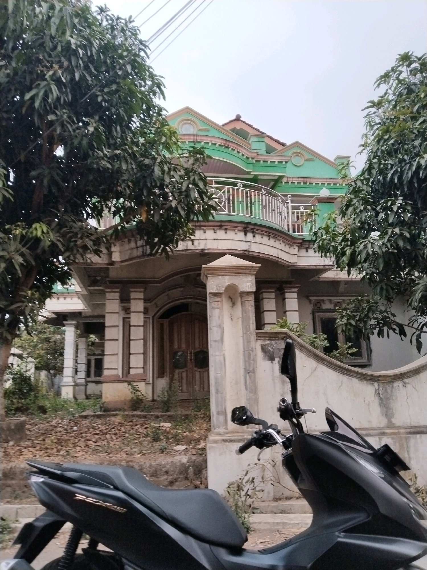 A motorcycle is parked in front of a two-story house in Sangkanjaya, Indonesia. The house has a green and cream facade with a prominent balcony on the second floor. Large trees with dense foliage flank the house. The foreground shows the back of a black scooter, with its seat and rear section visible. The ground is covered with dry leaves. The sky is overcast.