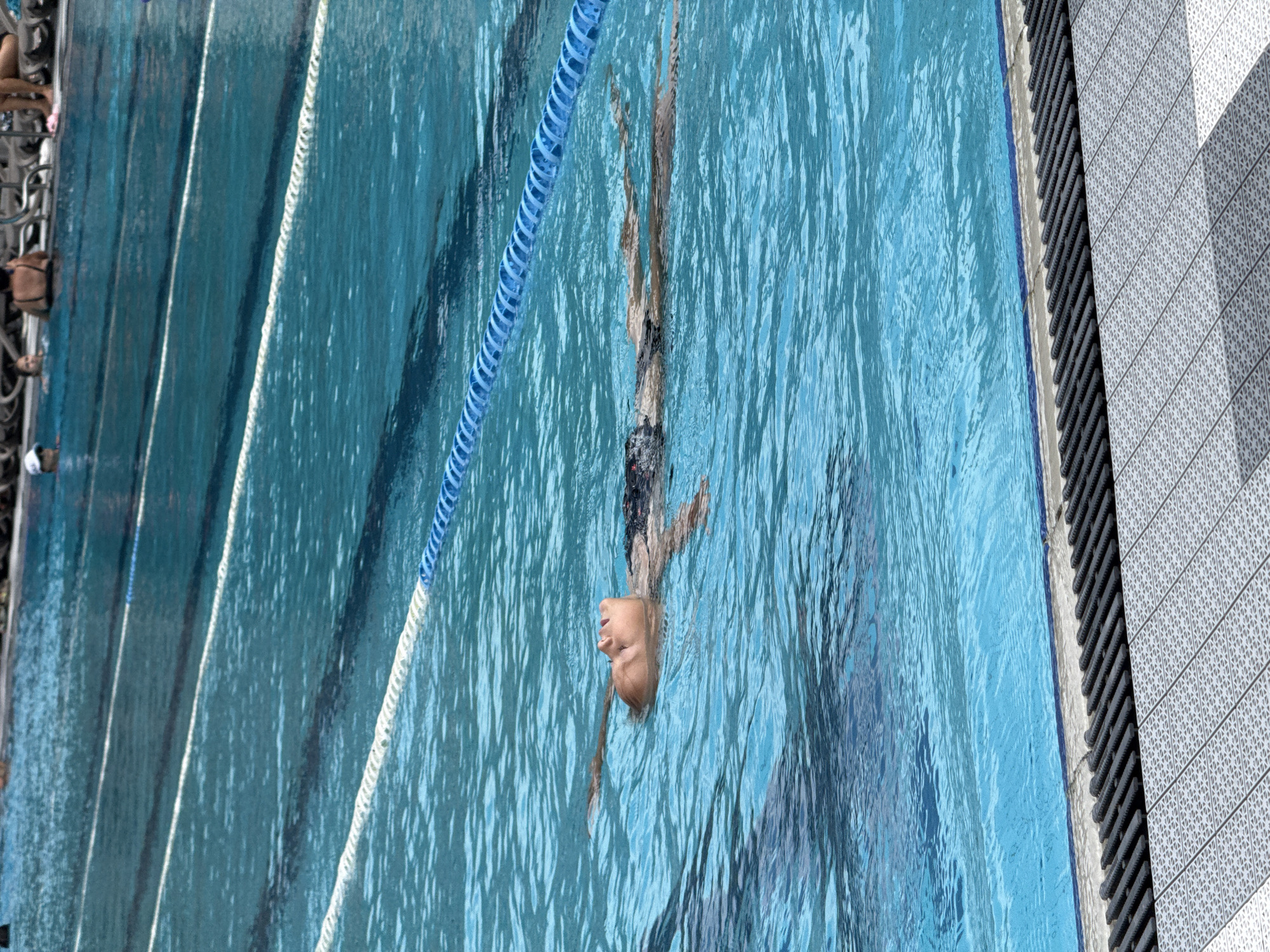 A child is floating on their back in a swimming pool, with their head tilted back and eyes closed. The water is a bright blue and there are lane ropes visible. The child is wearing a dark swimsuit. In the background, other people can be seen sitting on lounge chairs by the pool. The edge of the pool is visible, with a tiled walkway. The scene appears to be in a public pool.
