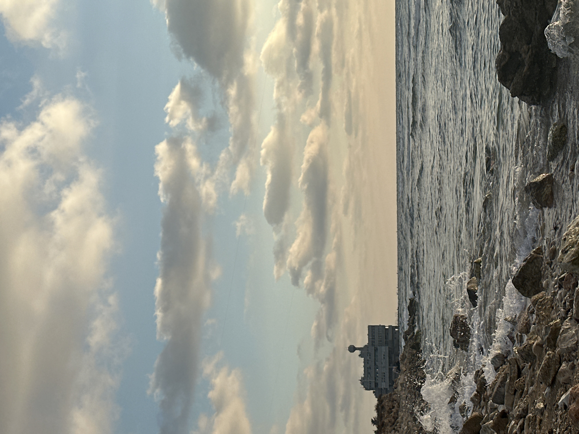 The image captures a coastal scene in Haifa, Israel, with a large, Brutalist-style building visible on the horizon, situated by the sea. The foreground is dominated by a rocky shoreline where waves crash, creating white foam and spray. The sea itself is choppy, with visible ripples and waves extending towards the horizon. The sky above is filled with scattered clouds, some appearing dense and dark, while others are illuminated by soft, warm light, suggesting either dawn or dusk. The building, which appears to be an industrial or institutional structure, has a distinctive, multi-story design with a rounded dome-like element on its roof. The overall impression is one of a stark, somewhat weathered coastal landscape.