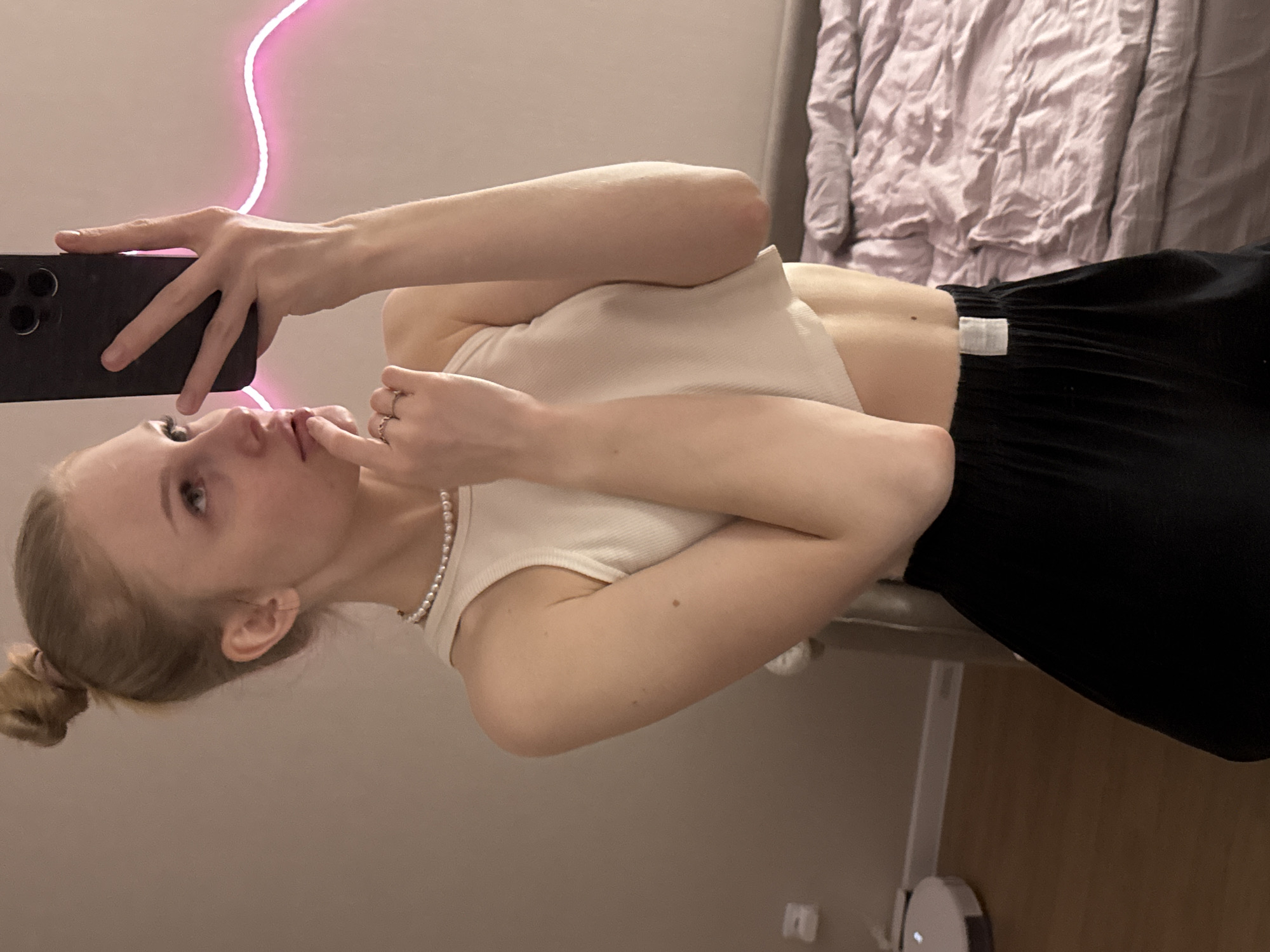 A young adult female with blonde hair tied in a high bun captures a mirror selfie indoors. She is seen from the waist up, wearing a white ribbed crop top, a pearl necklace, and black loose-fitting shorts. Her right hand, adorned with rings, holds a black smartphone with a multi-lens camera module, directed towards the mirror. Her left index finger rests gently on her lips as she gazes slightly upwards and to her right (her reflection's left). A subtle freckle is visible on her upper right arm and lower back. The background features a plain, light-colored wall with a decorative, undulating pink LED strip light running horizontally across it. To the right, a bed with a rumpled light purple duvet and a dark headboard is visible. The floor on the left consists of light wood planks, where a white circular robotic vacuum cleaner is situated. There are no discernible visual elements within the image to confirm the location as Perm, Russia.