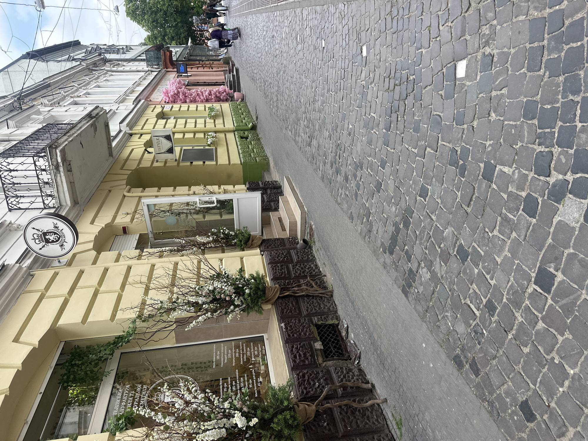 This image depicts a daytime scene on an urban street in the city of Ternopil, Ukraine, under overcast skies. The street is primarily paved with irregular dark grey and black cobblestones, bordered on the left by a smooth grey concrete sidewalk.
On the left side of the street, a multi-story building with a light yellow facade features distinct architectural elements including pilasters and cornices. At ground level, a commercial establishment identified by a sign in Ukrainian as "Lviv Soap and Natural Cosmetics Manufactory" occupies the space. A circular, dark sign bearing a crown and a crest is suspended above the primary entrance. The storefront includes two large display windows, with a white-framed glass door slightly ajar. Exterior decorations consist of several tall, artificial trees or bushes featuring white and pink flowers and bare branches, with burlap-wrapped bases resembling exposed roots. Green ivy cascades over the upper portion of the leftmost display window. The base of this building is constructed from dark brown, textured stone or concrete, containing a small, metal-grated vent. Further along this building, a black rectangular sign displaying a bra silhouette and the text "STYLE" is affixed, with two small white flower boxes positioned beneath a window.
Proceeding down the street, subsequent buildings include a white or cream-colored facade featuring a decorative black wrought-iron balcony on the second floor. Beyond this, another building's entrance is heavily adorned with extensive pink floral-like sculptural elements. Further right, a building with a darker facade displays a blue illuminated sign.
Approximately six to eight adult individuals are visible walking away from the camera in the mid-ground and background. One individual is clad in a dark floral skirt and a purple top, walking alongside another person. Another individual wears a white hooded sweatshirt. No specific interactions or activities beyond casual walking are discernible among the individuals.
Overhead electrical wires traverse the scene in the upper right quadrant. Green tree foliage is visible in the distant background on the right, indicating the presence of larger trees or a park area. The sky is uniformly light grey.