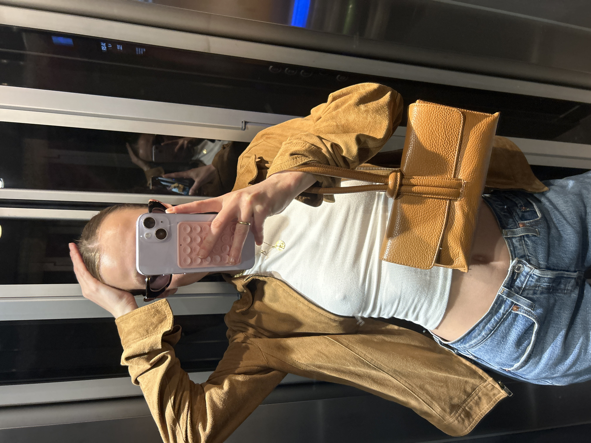 A fair-skinned person is taking a selfie with their phone in front of a reflective surface. The person is wearing a white ribbed top, a brown suede jacket, and light-wash blue jeans. A tan leather handbag hangs over their shoulder. The reflective surface shows a blurred image of the person holding the phone. The location appears to be indoors.