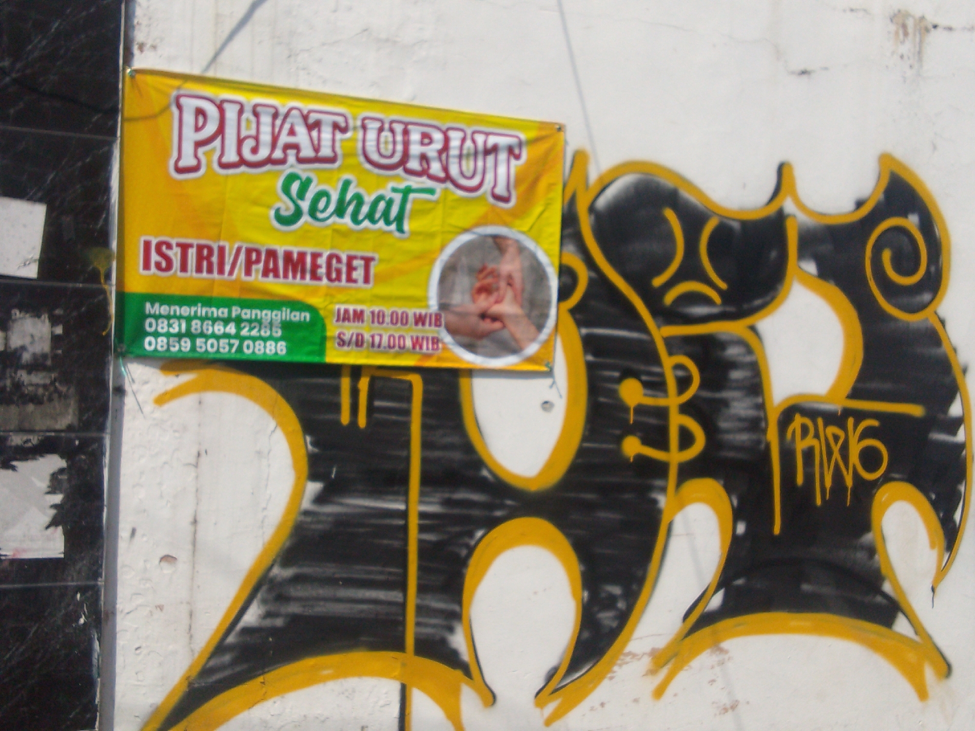 The image captures a close-up view of a white wall in Cianjur, Indonesia, adorned with a vibrant yellow banner and graffiti. The banner, positioned on the upper left of the wall, advertises "PIJAT URUT SEHAT" (Healthy Massage) and specifies that services are available for "ISTRI/PAMEGET" (Wives/Nannies). Contact information, including phone numbers "0831 8664 2285" and "0859 5057 0886", is clearly visible. Below this, opening hours are listed as "JAM 10.00 WIB S/D 17.00 WIB" (10:00 AM to 5:00 PM). A circular inset on the banner shows a close-up of hands giving a massage. The lower portion of the wall is covered in abstract graffiti featuring bold yellow outlines on a black background, with a tag that appears to read "PIF16" in yellow lettering. The wall itself shows signs of age and weathering, with some cracks and peeling paint visible.