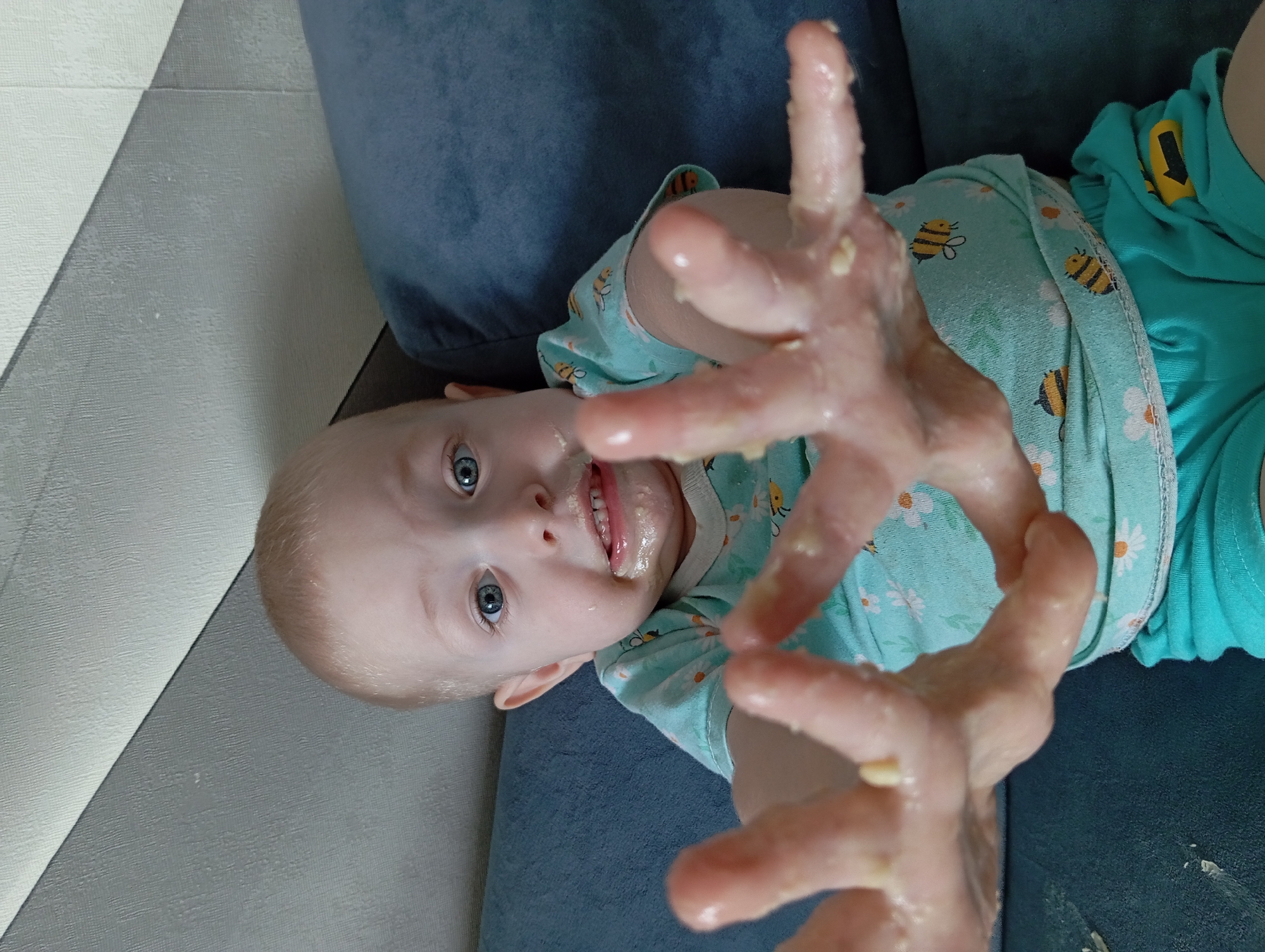 A toddler, lying on a dark blue surface that appears to be a couch, is the focal point of the image. The child is wearing a light blue patterned shirt and green shorts. The child's hands, covered in a wet, mushy substance, are extended towards the camera. Some of the same substance is on the child's face. The background includes a wall with a geometric design. The image was taken indoors, seemingly in a home or apartment, but there is no evidence to suggest the location of Yurgamysh, Russia.