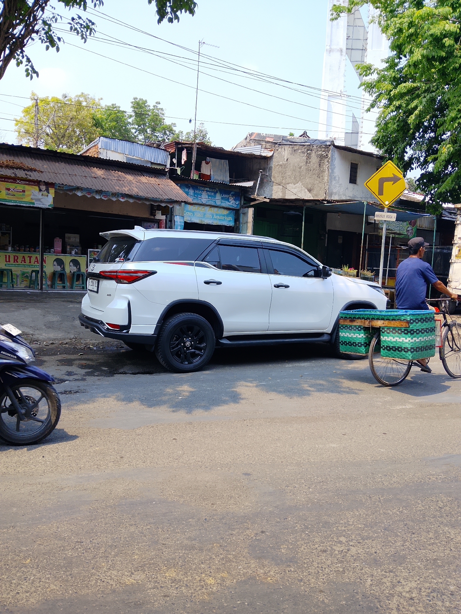 The image shows a white Toyota Fortuner parked on the side of a street in Ngelo, Indonesia. A blue Honda motorcycle is partially visible on the left side of the frame. A man wearing a blue shirt and a dark cap is riding a bicycle with a green woven basket attached to the back, moving away from the white SUV. The street is paved and shows signs of wear. In the background, there are several small shops with corrugated metal roofs. A yellow directional sign with a black arrow indicating a right turn is visible above the shops. Trees and utility poles with wires are also present. The sky is clear and blue.