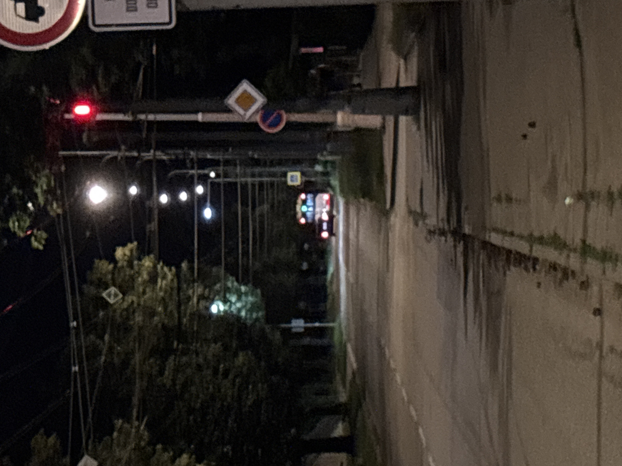 The image depicts a street scene at night in an urban environment, possibly in the city of Unknown, Unknown. A double-decker bus with illuminated windows is visible in the distance, moving along the road. The road is paved with white lane markings. On the right side of the road, a sidewalk is present, lined with trees and streetlights. A traffic signal with a red light illuminated is prominently displayed on a pole. Several road signs are also visible on this pole, including a diamond-shaped sign with a yellow inner square and a circular sign with a red border and a blue inner circle with a red diagonal line. The background consists of trees and the night sky, with overhead power lines visible. The lighting suggests it is nighttime, with artificial lights from streetlamps and vehicles illuminating the scene.