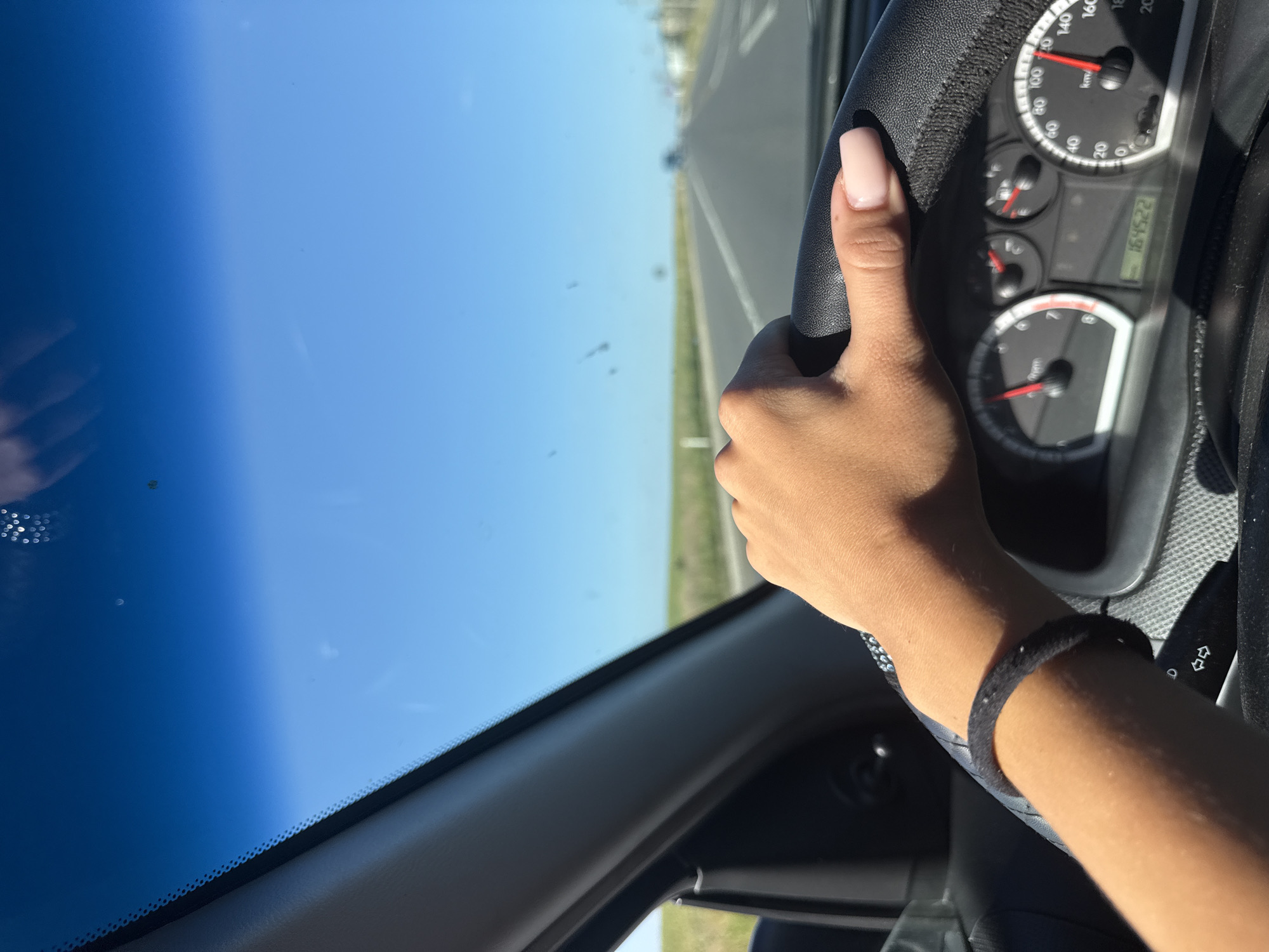 The image shows a first-person perspective of a person driving a car on a sunny day. The driver's hand, with light pink fingernails, is resting on the steering wheel. A black bracelet is visible on the driver's wrist. The car's dashboard is in the foreground, with the speedometer indicating a speed of approximately 100 km/h and the odometer showing 164522. The windshield is clear, revealing a bright blue sky and a flat, green landscape with a straight road extending into the distance. The scene suggests the car is traveling on a highway outside of a populated area, possibly in the countryside. No specific landmarks of Comrat, Moldova are visible.