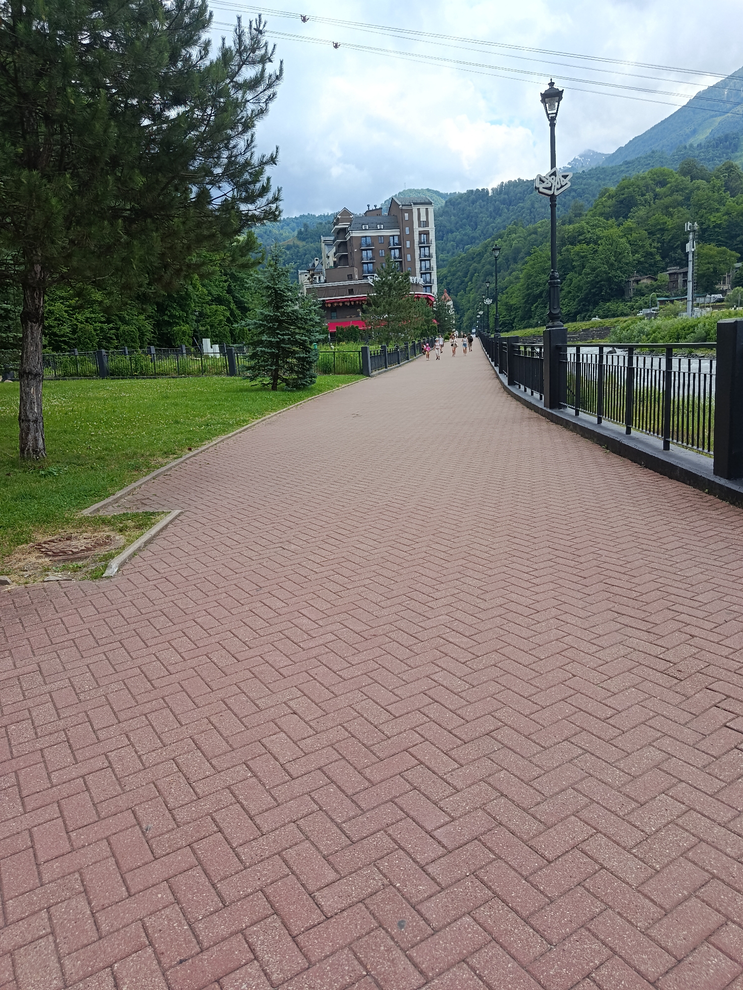 The image shows a scenic view of a paved walkway in Estosadok, Russia, likely along a river or body of water. The walkway is constructed with red brick pavers laid in a herringbone pattern and extends into the distance, curving slightly to the right. Along the left side of the walkway, there is a grassy area with trees and a black metal fence. In the background, a multi-story building with a modern design is visible, nestled among lush green trees on a mountainside. A river flows along the right side of the walkway, separated by a black metal railing. Several people are walking on the path in the middle distance, appearing to be tourists enjoying the scenery. The sky is partly cloudy, with hints of blue. The overall impression is one of a well-maintained recreational area in a mountainous resort town.
