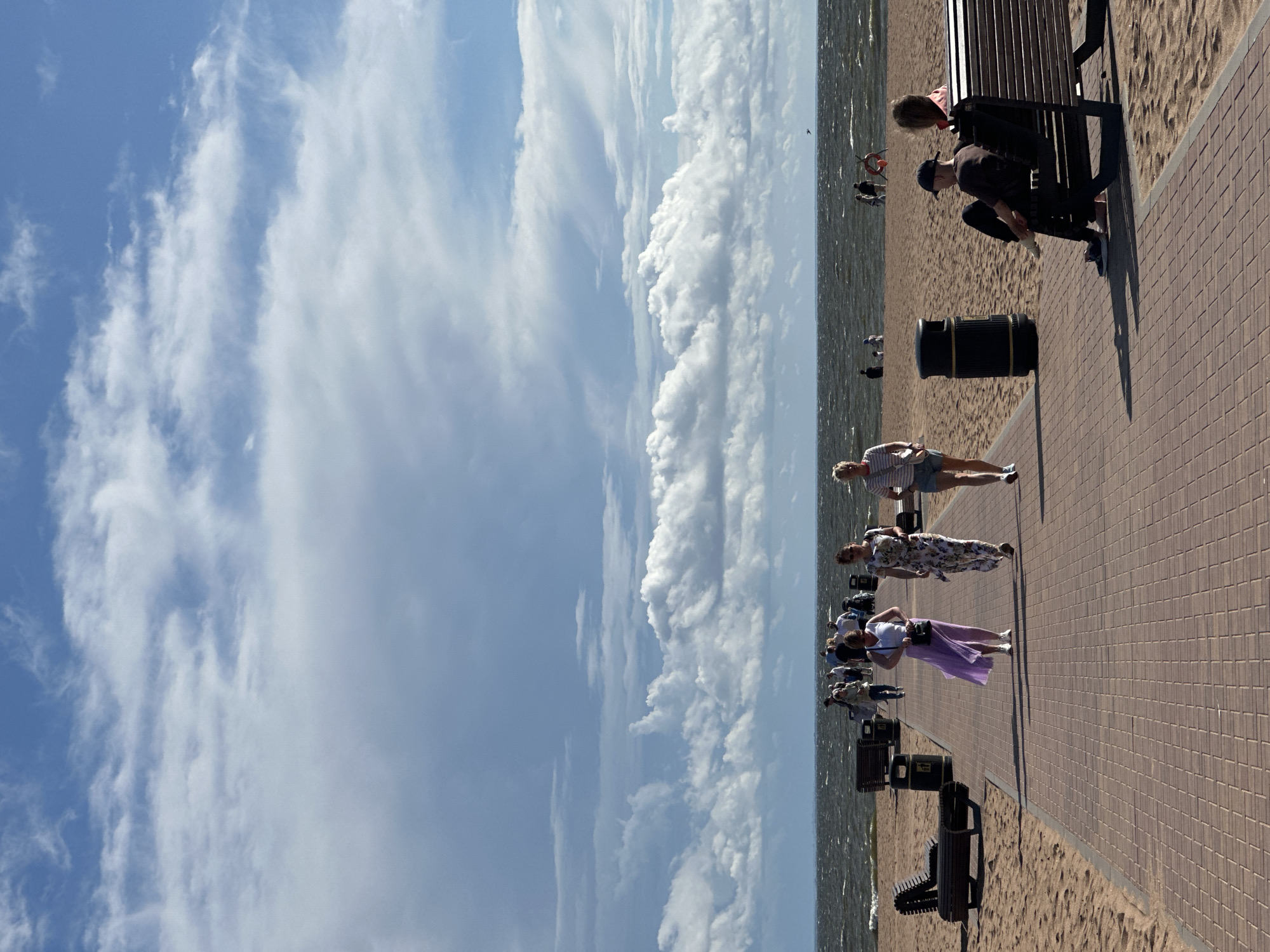 The image depicts a sunny day at a beach in Zelenogorsk, Russia. A paved walkway runs parallel to the sandy beach, with benches and trash cans lining its edge. Several people are strolling along the walkway, while others are seated on the benches, observing the sea. The sea itself is active, with moderate waves crashing onto the shore. The sky is a mix of blue and white clouds, suggesting a partly cloudy day. The overall atmosphere is one of leisure and recreation, with individuals enjoying the seaside environment.