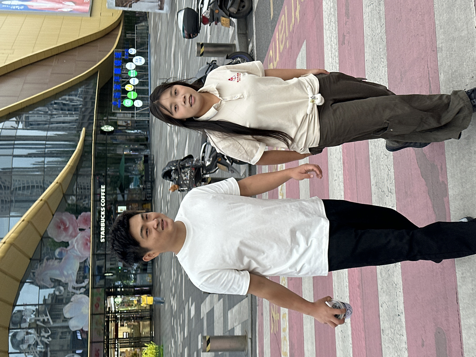 A young Asian man and a girl are standing on a pink and white striped crosswalk in Jiangjin, China. The man is wearing a white t-shirt and black pants and holding a small container in his right hand. The girl is wearing a light-colored polo shirt and dark green pants. In the background is a building with a Starbucks Coffee shop on the ground floor. Several scooters are parked nearby. The street is paved with grey tiles, and there are metal bollards along the crosswalk.