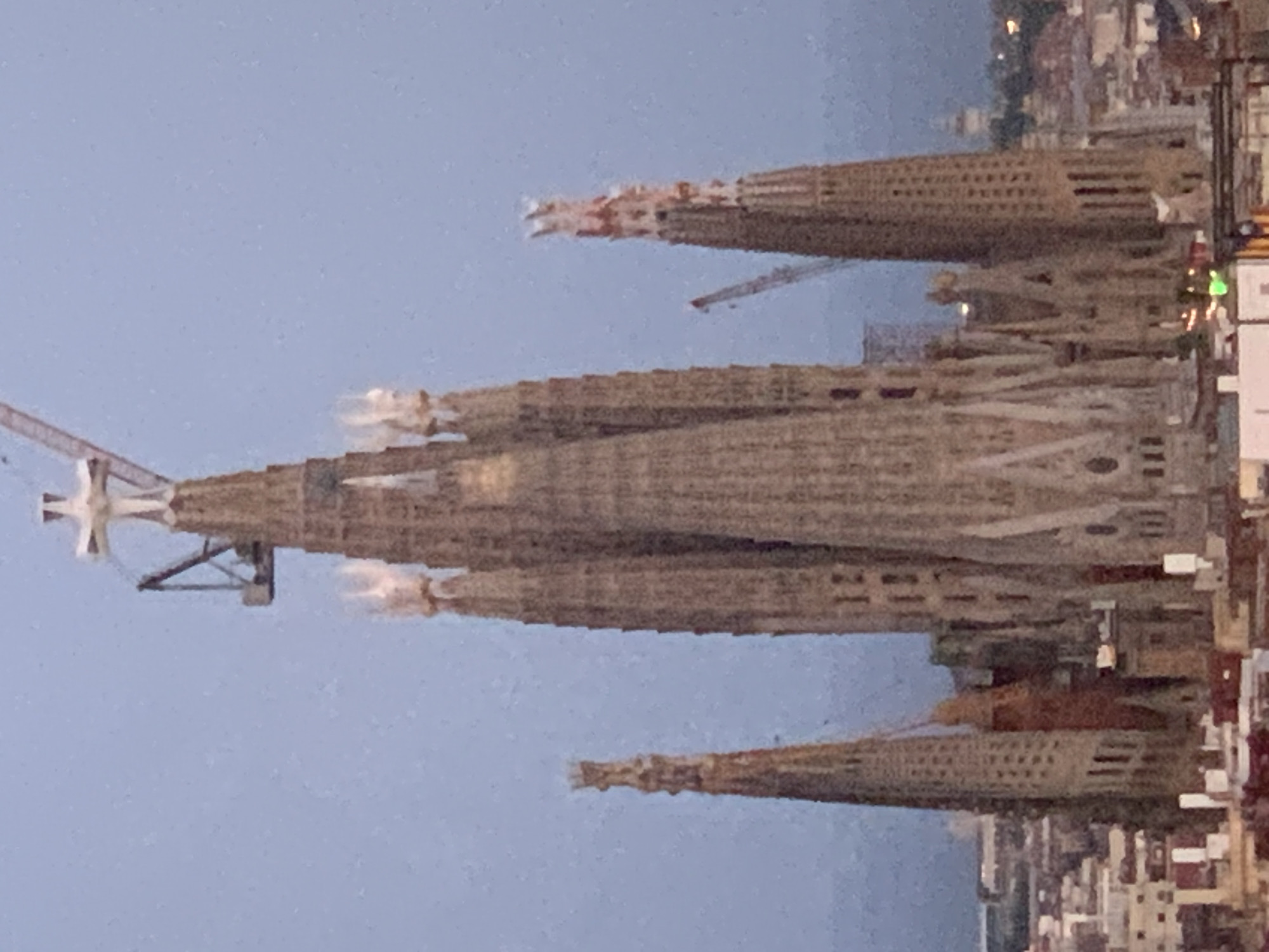 The image is a close-up, aerial view of the Sagrada Familia in Barcelona, Spain. The iconic basilica, under construction, dominates the frame with its towering spires and intricate facades. Several cranes are visible, indicating ongoing work on the structure. The sky is a muted blue, suggesting either early morning or late evening. In the foreground, the rooftops of surrounding buildings in Barcelona are visible, a mix of apartments and older structures, with some lights appearing in the distance. The lighting is soft, and the overall impression is of a grand architectural masterpiece viewed from above.