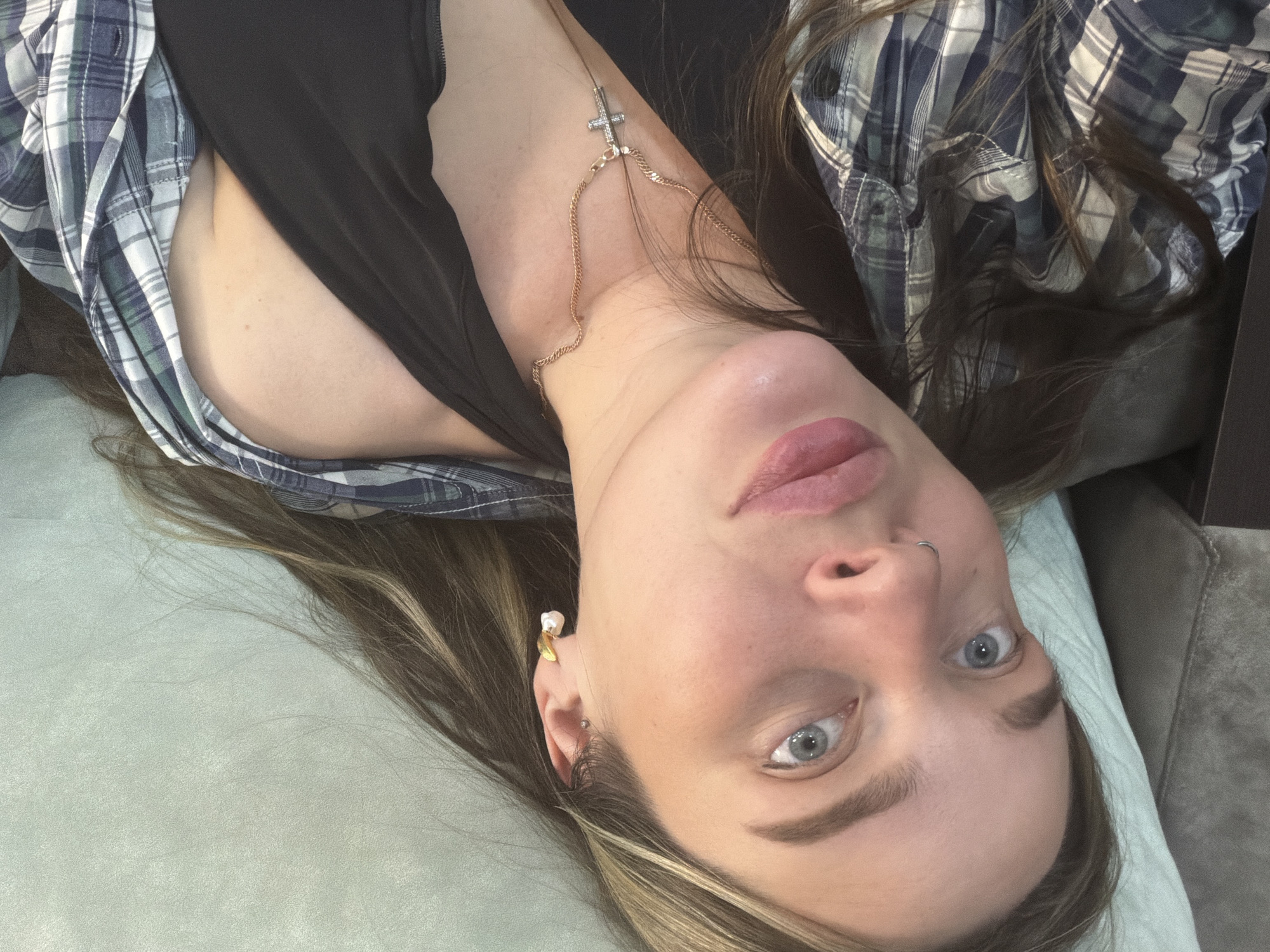 A young woman is captured in an intimate, close-up indoor shot, lying down and looking directly into the camera. The image's perspective is slightly inverted due to her head position. She has fair skin, wide-open light blue eyes, and full, pink-tinted lips. Her eyebrows are well-defined, and she has a silver hoop nose ring in her left nostril. Her long, brown hair with blonde highlights is spread out around her head and visible on the light greenish-blue upholstered surface she is lying on, which could be a bed or couch.
She is dressed in a dark, possibly black, tank top or camisole, worn beneath an open plaid shirt featuring shades of blue, white, and dark green. A delicate gold chain with a small, sparkly cross pendant is draped across her chest and shoulder. In her right ear, she wears a decorative earring, appearing to be a gold element with a white, pearl-like adornment, along with another small ear piercing. A darker grey or brown upholstered surface is partially visible in the background, to her right. Her expression is neutral and direct, conveying a sense of alertness. The setting is an indoor space in Chaikovsky, Russia. There is no visible text in the image.