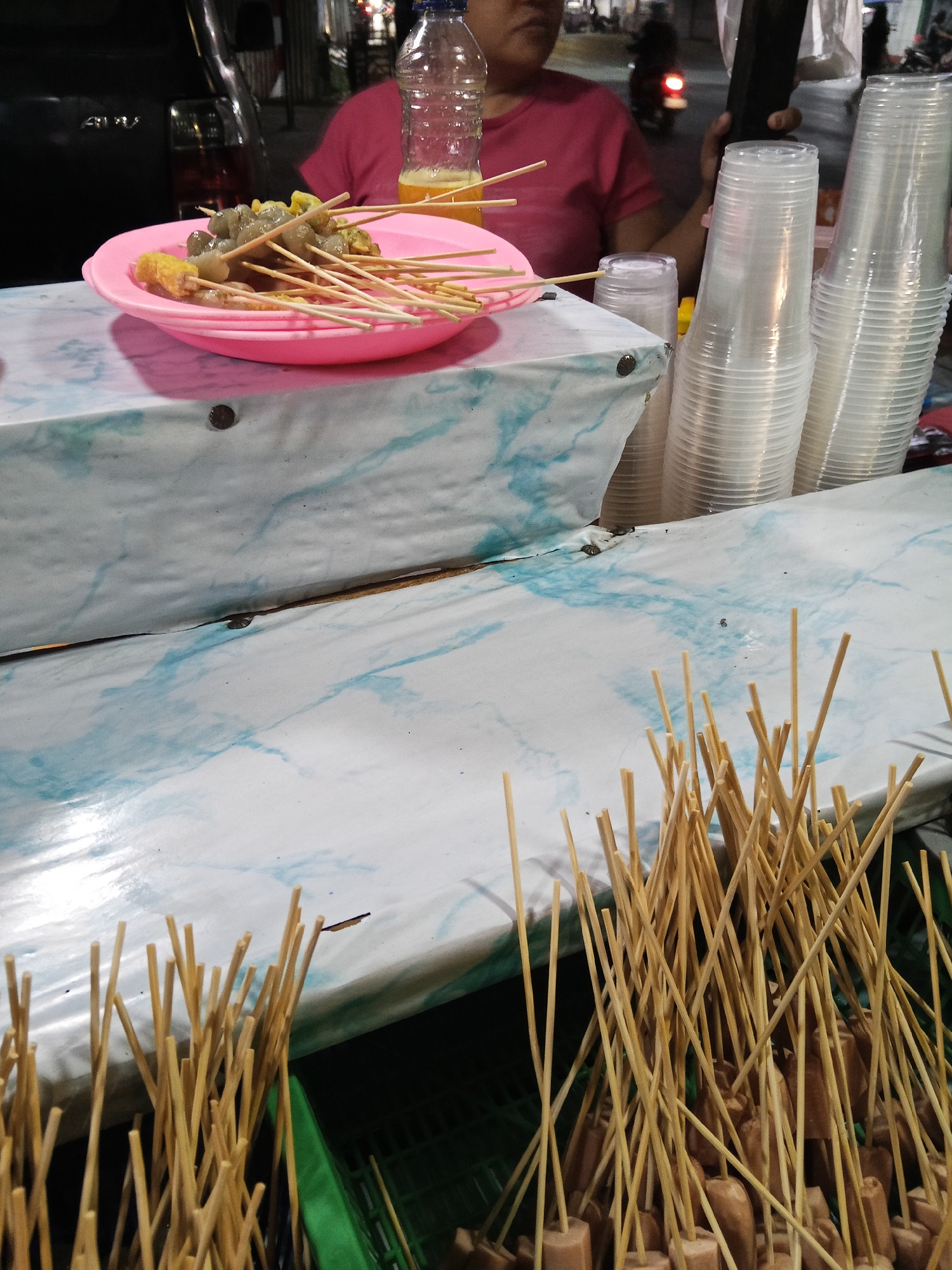 This is a close-up, eye-level shot of a street food stall at night in Lubuklinggau, Indonesia. In the foreground, numerous wooden skewers are arranged vertically, some with what appear to be sausages or meatball pieces. A pink plastic plate filled with skewers of meatballs and fried tofu sits on a table covered with a white and blue marbled fabric. Behind the plate, a person wearing a pink t-shirt is partially visible, holding a mobile phone. Stacked plastic cups are positioned to the right of the plate, and a glass of dark liquid and a bottle of clear liquid with yellow contents are also present on the table. The background is dark, with faint lights suggesting a city environment.