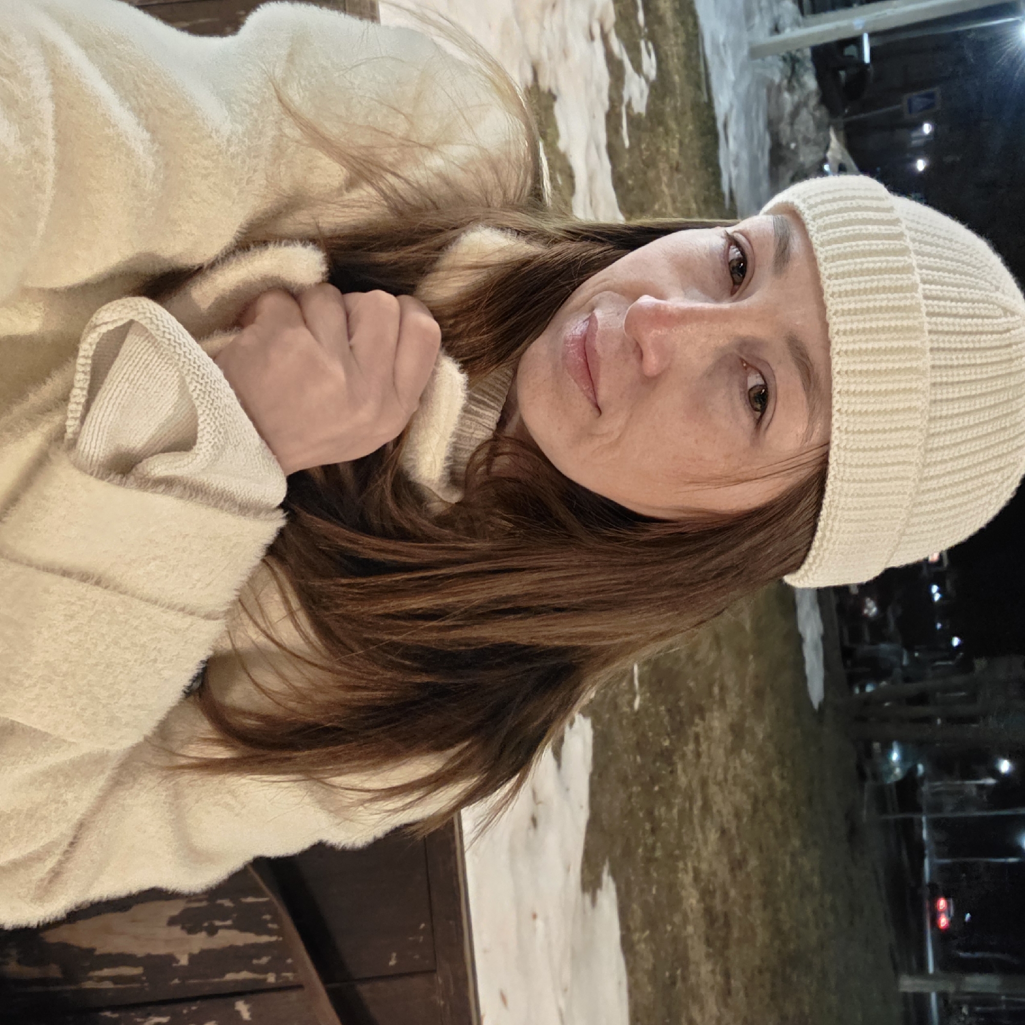 A female subject is depicted seated outdoors at night in Chaikovsky, Russia. She wears a cream-colored ribbed beanie, a light-colored fuzzy coat over a similar-toned ribbed sweater, and has long brown hair. Her hands are clasped together at chest level, holding the front of her coat. She has brown eyes and is looking directly forward. The background shows a street scene illuminated by multiple streetlights. Patches of snow and brown grass are visible on the ground, indicating a cold season. In the distance, several vehicles are present, including one with illuminated red taillights and another with faint headlights. A dark vehicle is partially visible to the left of the subject, near a utility pole. A diamond-shaped road sign with a white background and black symbol is also visible in the upper left background. The subject is seated on a dark wooden bench, partially visible on the right.