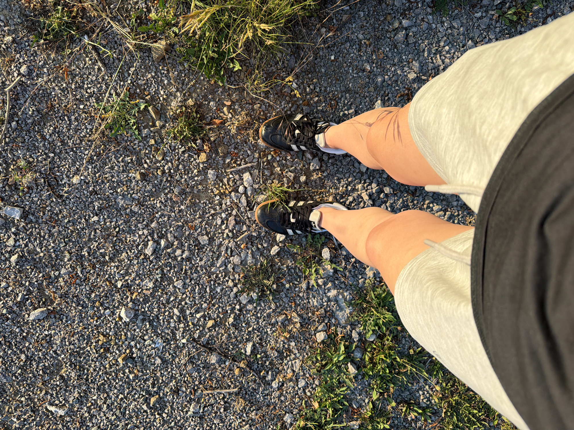 The image shows a top-down view of a person's legs and feet standing on a gravel path. The person is wearing black athletic shoes with white accents and light-colored shorts. Patches of green grass and sparse weeds are interspersed with the gravel. The lighting suggests it is either early morning or late afternoon, casting shadows on the ground. The background is out of focus and appears to be more gravel and vegetation.