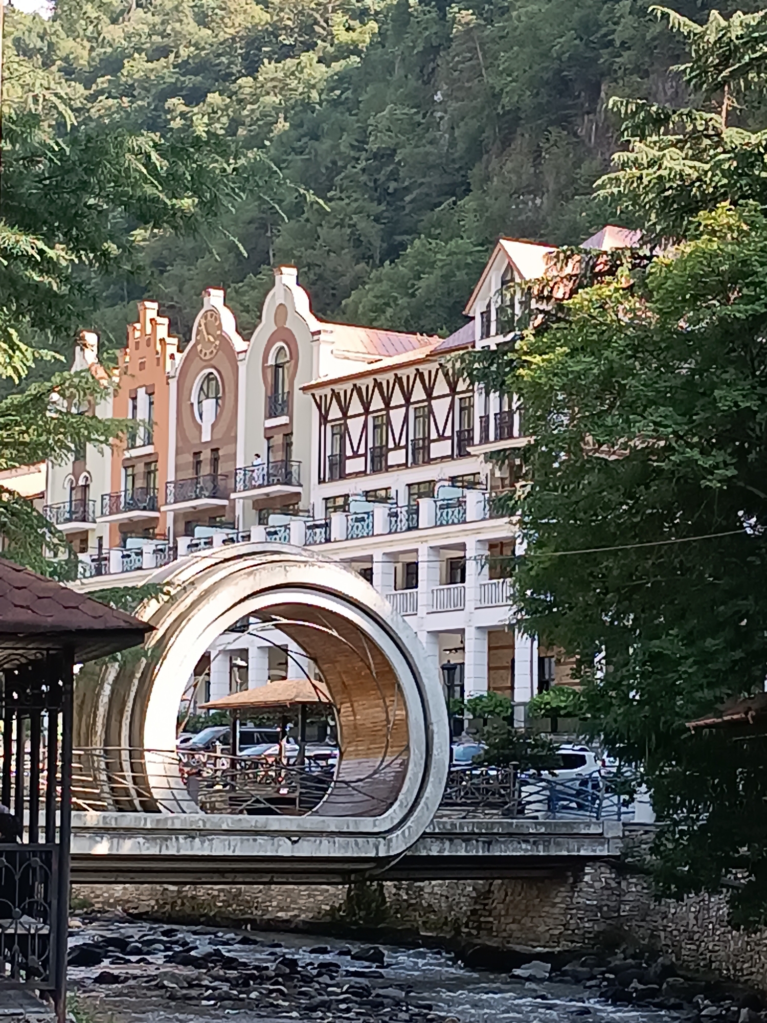 The image captures a view of the "Central Park Bridge" in Borjomi, Georgia. The bridge features a distinctive large, circular concrete structure resembling a tunnel, with a wooden-like interior, allowing passage over a rocky riverbed. Beyond the bridge, a multi-story building with a white facade, ornate balconies, and distinctive Dutch gable roofs is visible, set against a backdrop of lush green trees and a mountainous forest. The scene is illuminated by daylight, suggesting it was taken in the daytime. There are no visible people in the foreground or mid-ground, but a few cars are visible through the circular opening of the bridge. The architecture of the building suggests a historical or European influence.