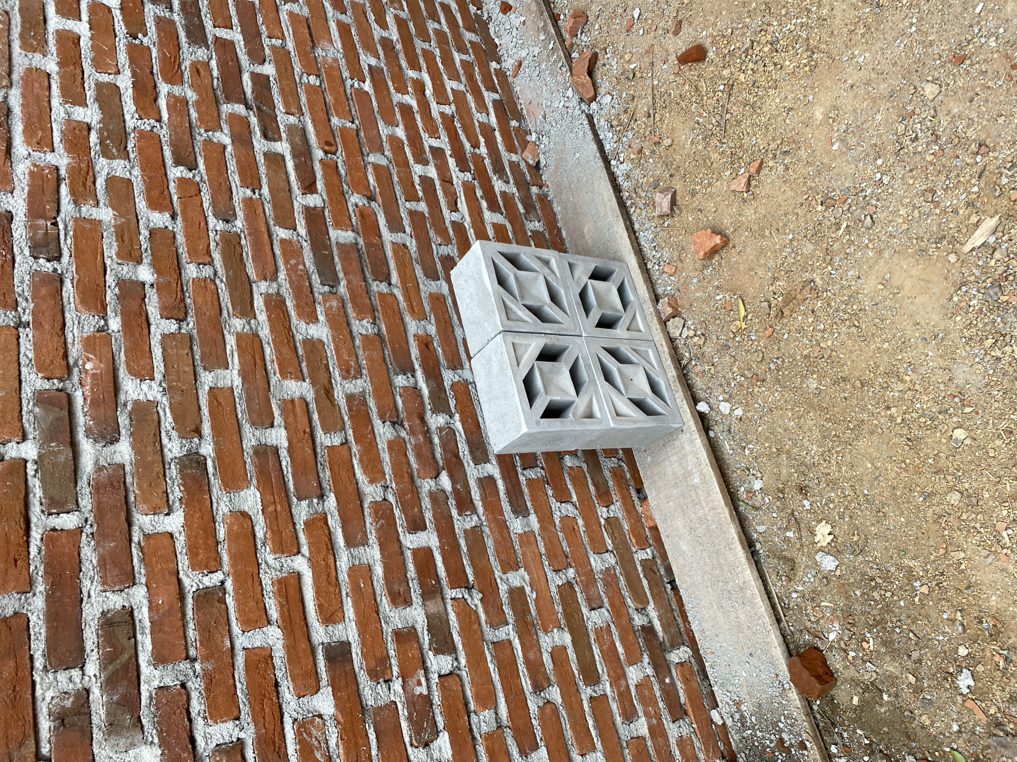 The image depicts a construction site with a partially constructed red brick wall in Banda Aceh, Indonesia. Two grey concrete blocks with decorative cutouts are placed in front of the wall, resting on a wooden plank. The ground is covered with dirt, small stones, and scattered brick fragments, indicating ongoing construction activity. The wall itself appears to be laid with mortar, and the overall scene suggests an outdoor setting.