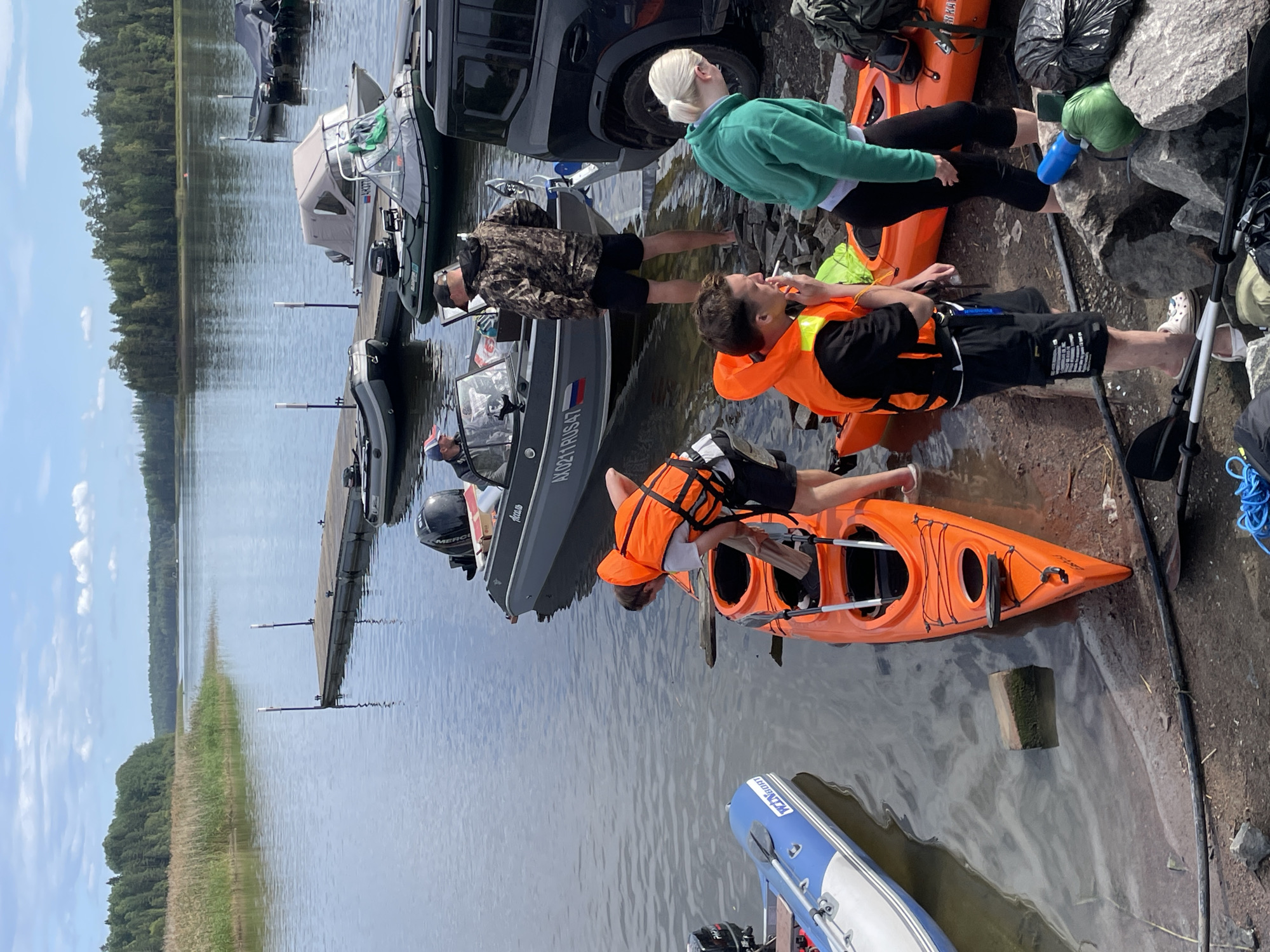 In Berozovo, Russia, on a sunny day with a partly cloudy sky, a group of people are gathered near the water's edge. The scene is set on the bank of a wide river or lake, with a wooden dock extending into the water and several boats moored nearby. Two orange kayaks are positioned on the shore.
A young man wearing an orange life vest and black shorts is seen leaning over one of the kayaks, holding a wooden plank. He is smoking a cigarette. Beside him, a young woman in a green hoodie and black leggings is also near the kayaks. Another man in a camouflage jacket and black shorts stands near a speed boat, looking towards the water.
In the background, a large grey speed boat with the registration "AK0211RUS47" is visible. Several other boats, including an inflatable one and a larger covered vessel, are docked or anchored in the water. The riverbank is rocky and uneven, with some vegetation and tall reeds on the distant shore, backed by a dense forest. The overall atmosphere suggests preparation for or a break from water activities.