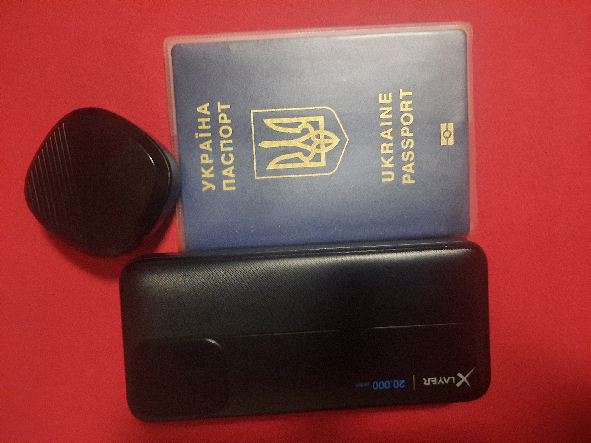 The image shows a close-up, overhead view of a dark red surface with several items placed on it. From left to right, there is a black portable charger with "X LAYER 20.000 mAh" printed vertically on its side. To the right of the charger is a blue Ukrainian passport, showing the text "УКРАЇНА ПАСПОРТ" and "UKRAINE PASSPORT" in gold lettering, along with a gold emblem. Above the passport and slightly to the right is a small, black, irregularly shaped object with a ribbed texture on its top surface. The lighting suggests an indoor setting, possibly artificial. There are no people or animals present in the image. The background is a plain red surface. The image does not provide any information or context that would allow for the determination of its location in Rosenheim, Germany.