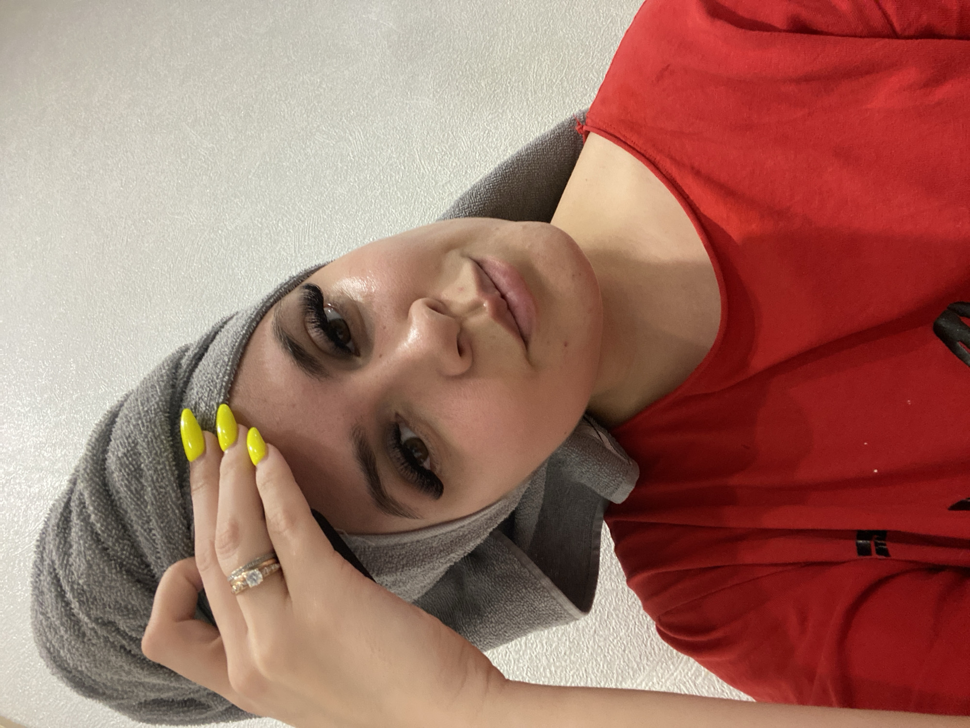 A woman with light skin, long eyelashes, and yellow fingernails, poses with a gray towel wrapped around her head. She is wearing a red t-shirt. A ring is visible on her left hand. The background is a white, textured wall. The location is potentially in the city of Chaikovsky, Russia.