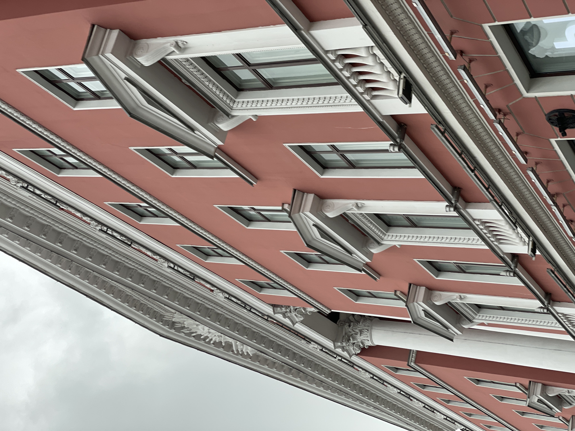 The image depicts a section of a building in Saint Petersburg, Russia, characterized by its red exterior and white architectural details. The view is angled upwards, capturing multiple windows with white frames and decorative elements. Ornamental molding runs along the building's edge. The sky is overcast. There are no people visible in the scene.